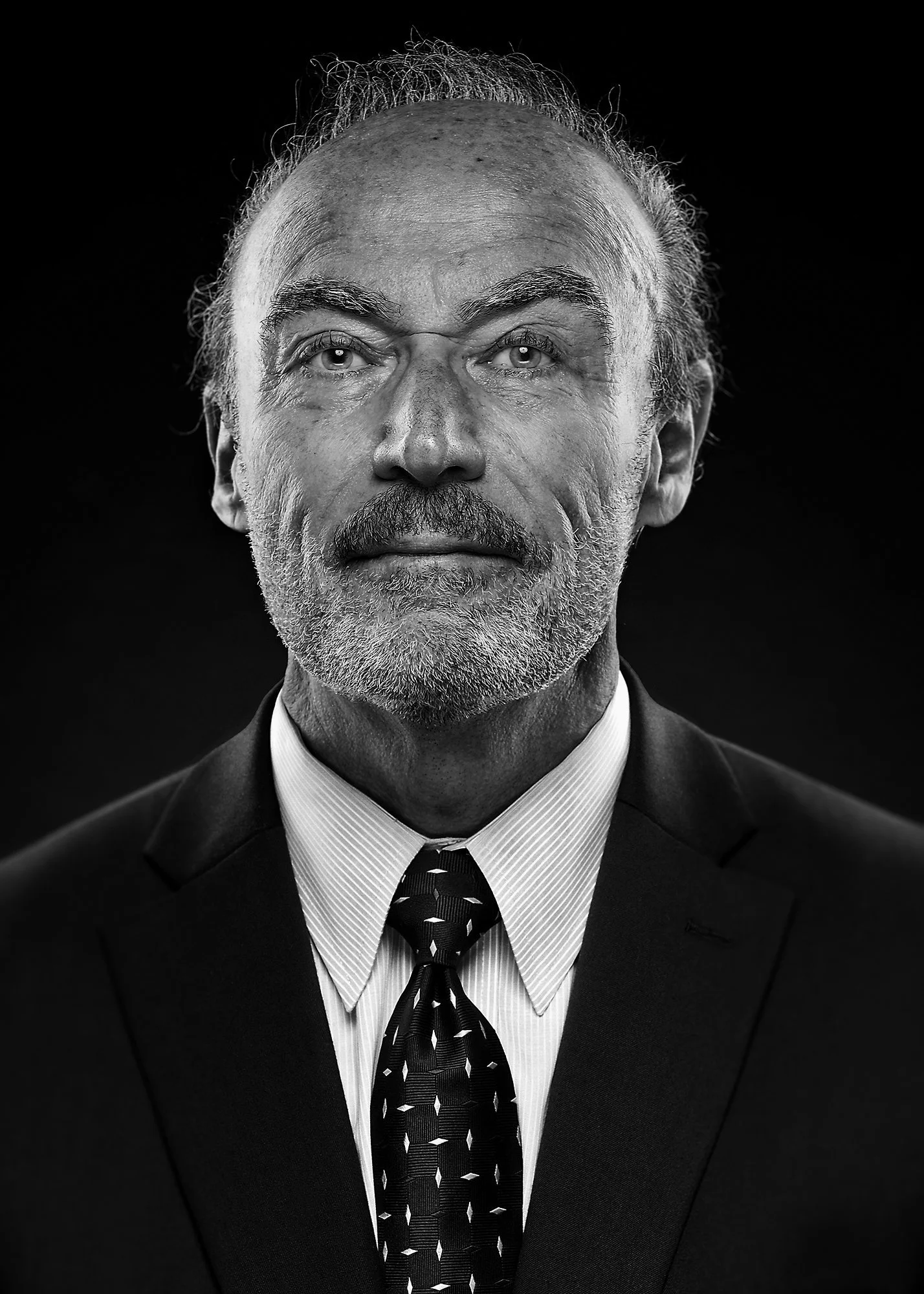 Black-and-white portrait of a middle-aged man with gray hair and beard, wearing a suit, white shirt, and patterned tie, against a dark background.