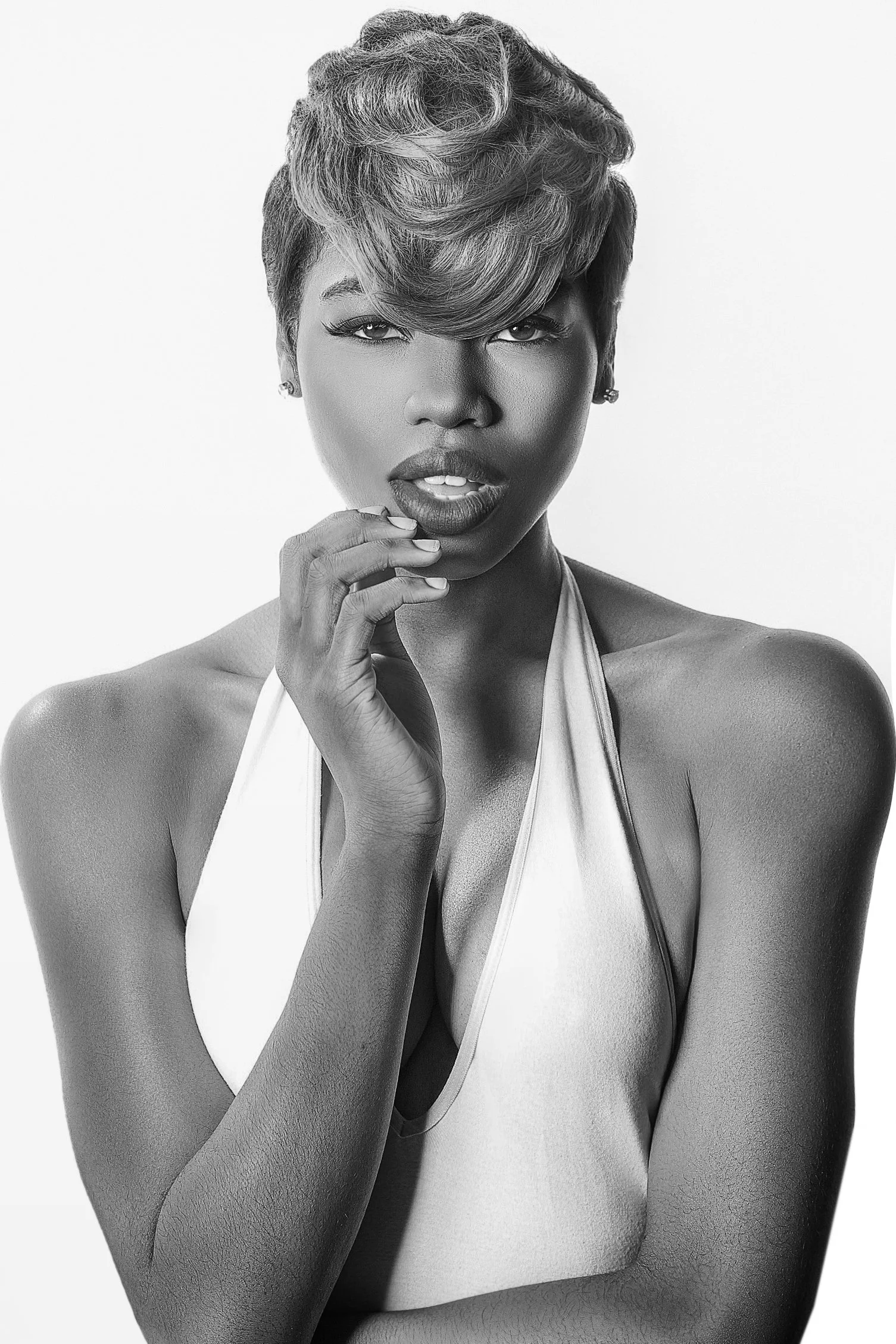 A black and white portrait of a woman with short, styled hair, wearing a halter top, posing with her hand near her face. She has a confident expression and is looking directly at the camera. Fashion Photography www.exvotostudio.com Best-Fashion-Portr