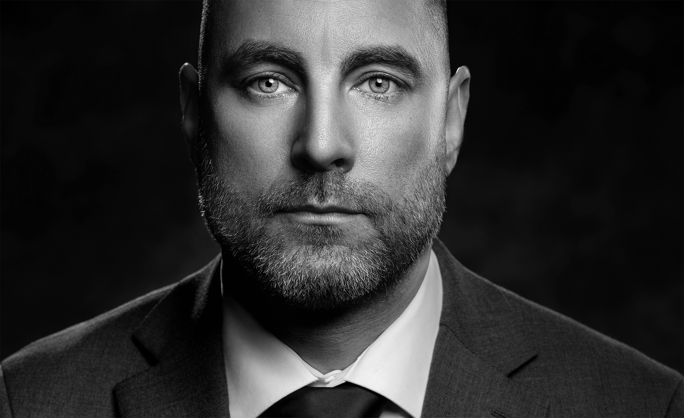 Black and white close-up portrait of a man in a suit with a beard and short hair, looking directly at the camera. In office headshot and branding photographer in Maryland, PA, VA and Washington D.C. by www.exvotostudio.com
