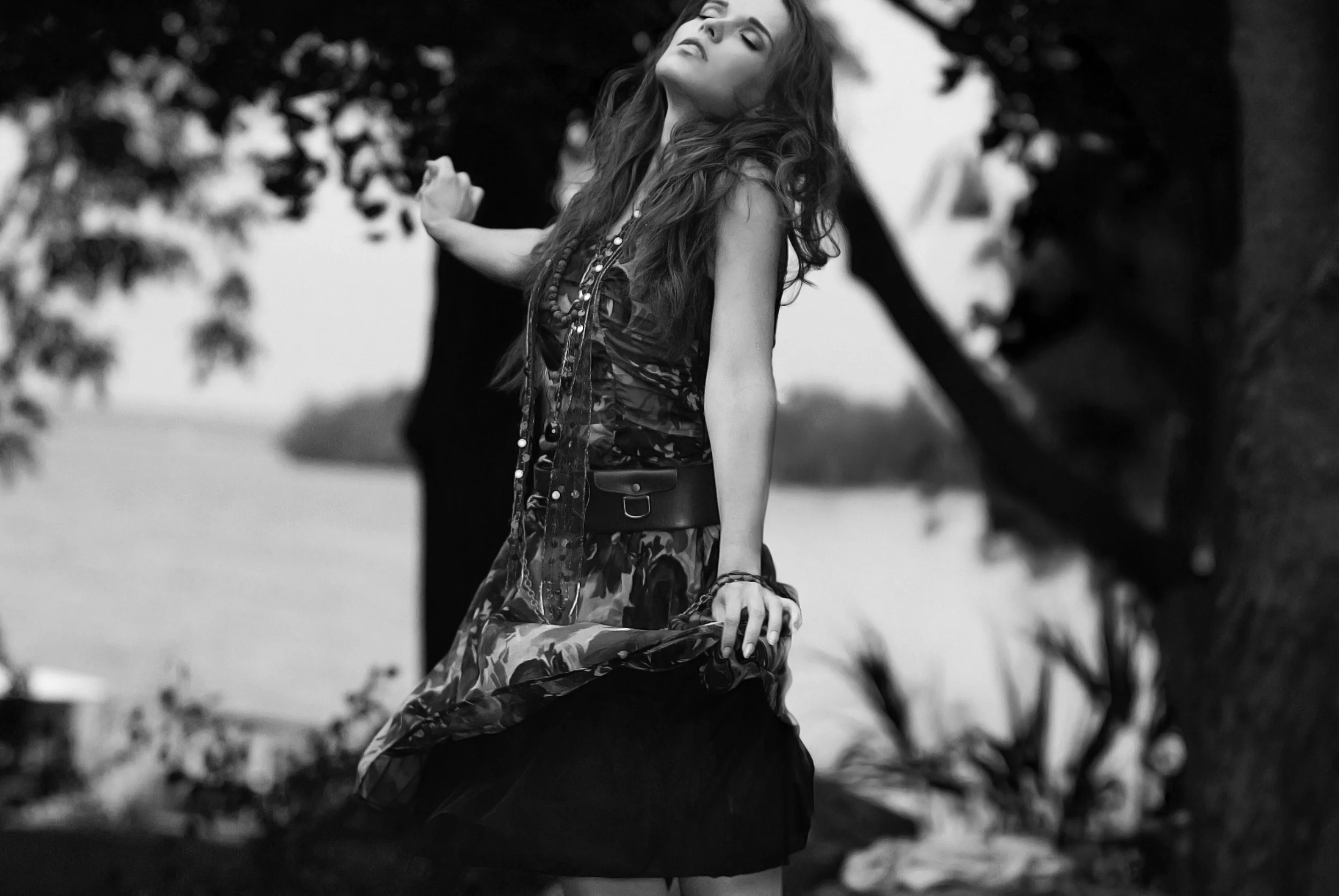 A woman with long wavy hair wearing a patterned dress and jewelry, standing outdoors near a tree with water in the background, captured in black and white. Fashion Photography www.exvotostudio.com