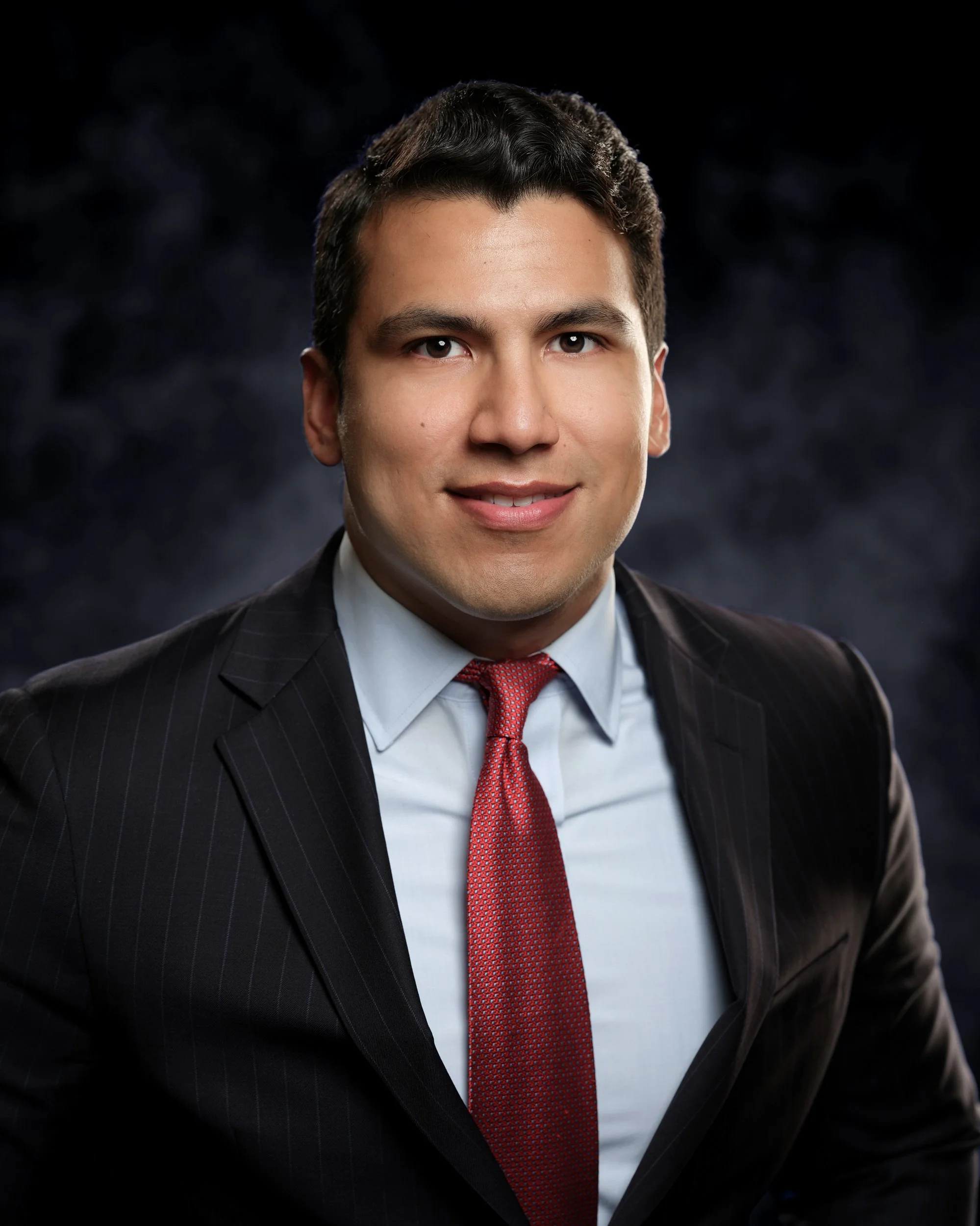 Professional headshot of a young man in a black suit with pinstripes, white shirt, red tie, with dark hair, smiling, against a dark, textured background.