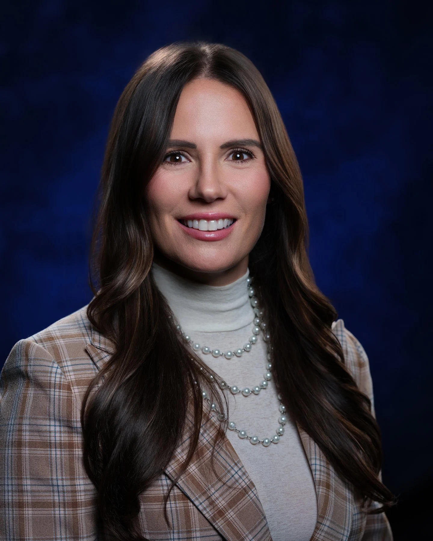 Professional portrait of a woman with long wavy brown hair wearing a beige plaid blazer, white turtleneck, and layered pearl necklaces, smiling against a dark blue background. In office headshot and branding photographer in Maryland, PA, VA and Washi