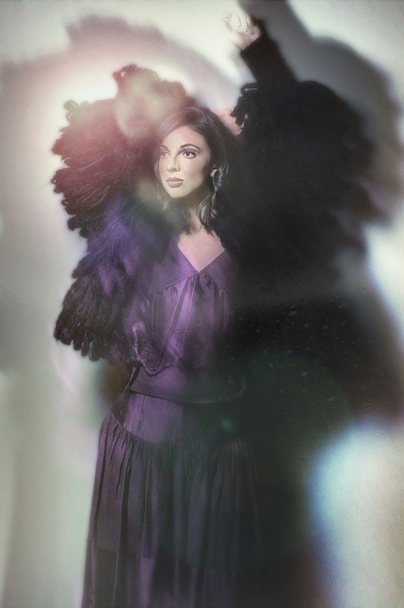 A woman with light skin and dark hair wearing a purple dress and a large black fur-like coat, posed with her left arm raised and head turned slightly to the right, with colorful light effects around her. Best-Fashion-Portrait-Commercial-Business-Head