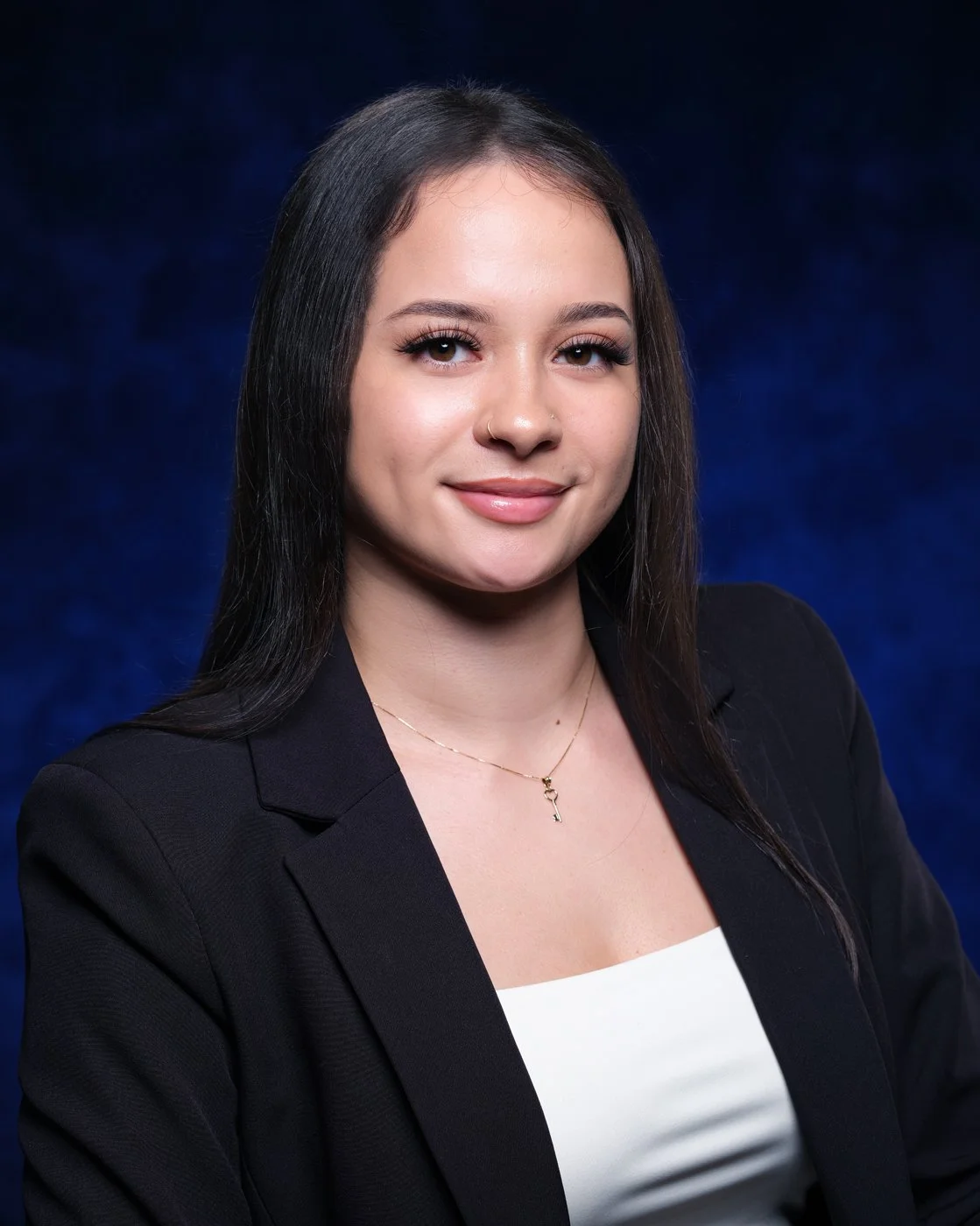 Professional portrait of a young woman with dark hair, wearing a black blazer and white top, against a dark blue background.