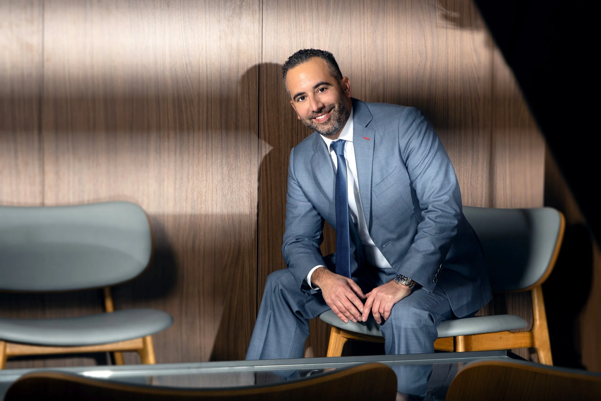 A man in a light blue suit and white shirt sitting on a chair in front of wooden paneling, smiling and looking at the camera.