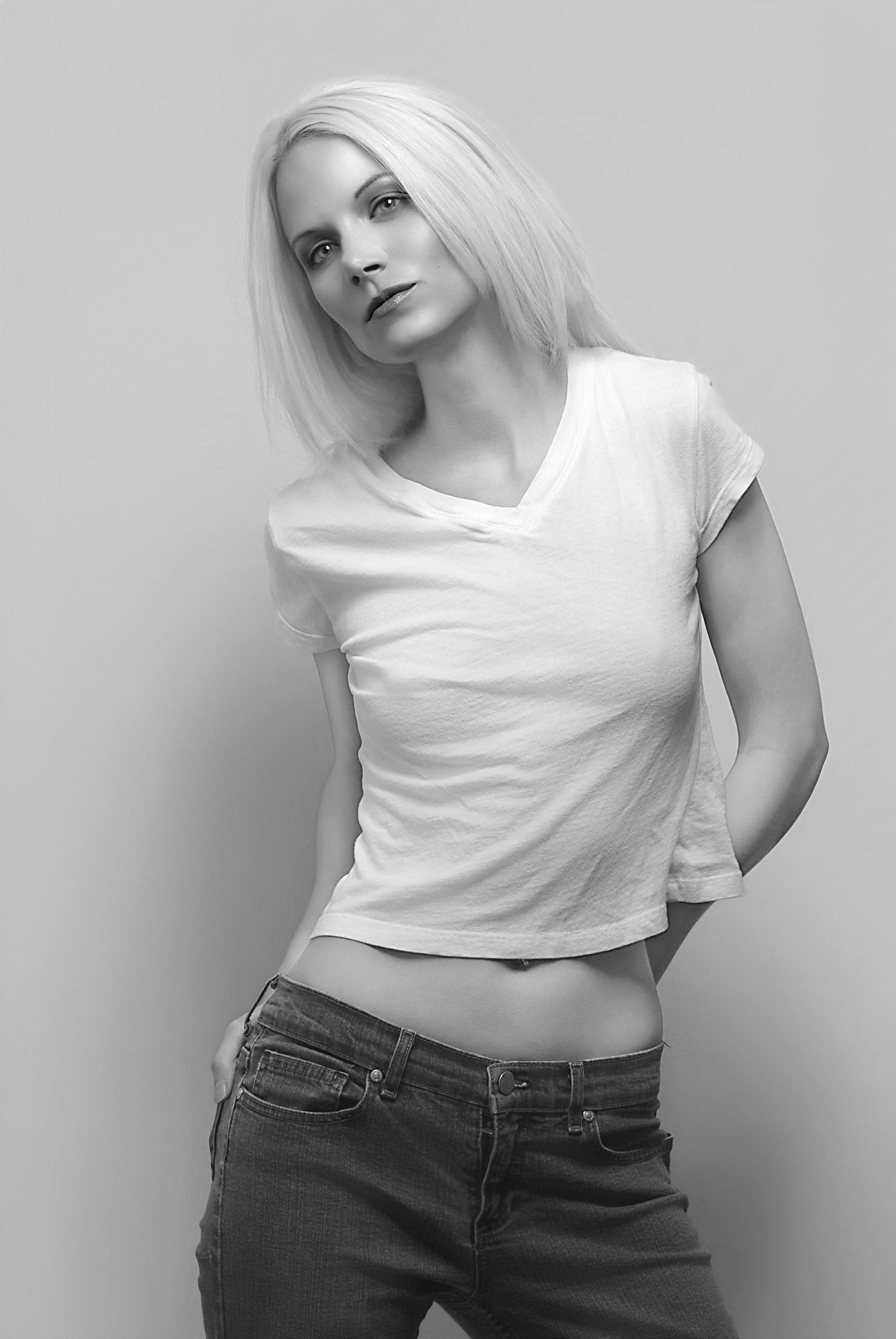 Black-and-white photo of a young woman with shoulder-length blonde hair, wearing a white t-shirt and dark jeans, standing against a plain background with her arms behind her back, leaning slightly to one side. Best-Fashion-Portrait-Commercial-Busines