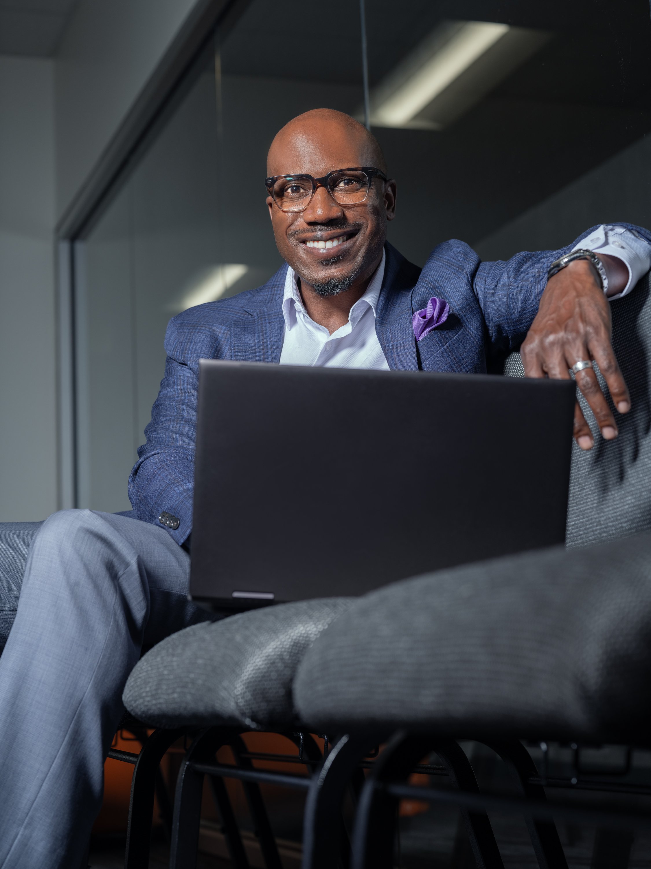 A smiling man in a blue blazer and glasses sitting on a gray chair with an open laptop in front of him, in a modern office setting.