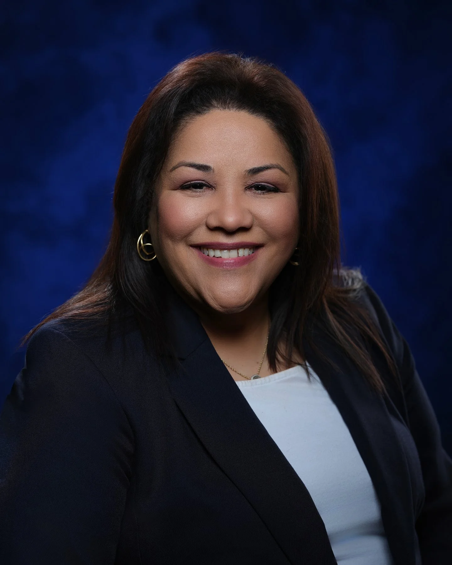 A professional woman with dark brown hair, smiling, wearing a navy blazer, white top, gold hoop earrings, and a delicate necklace, against a dark blue background. In office headshot and branding photographer in Maryland, PA, VA and Washington D.C. by