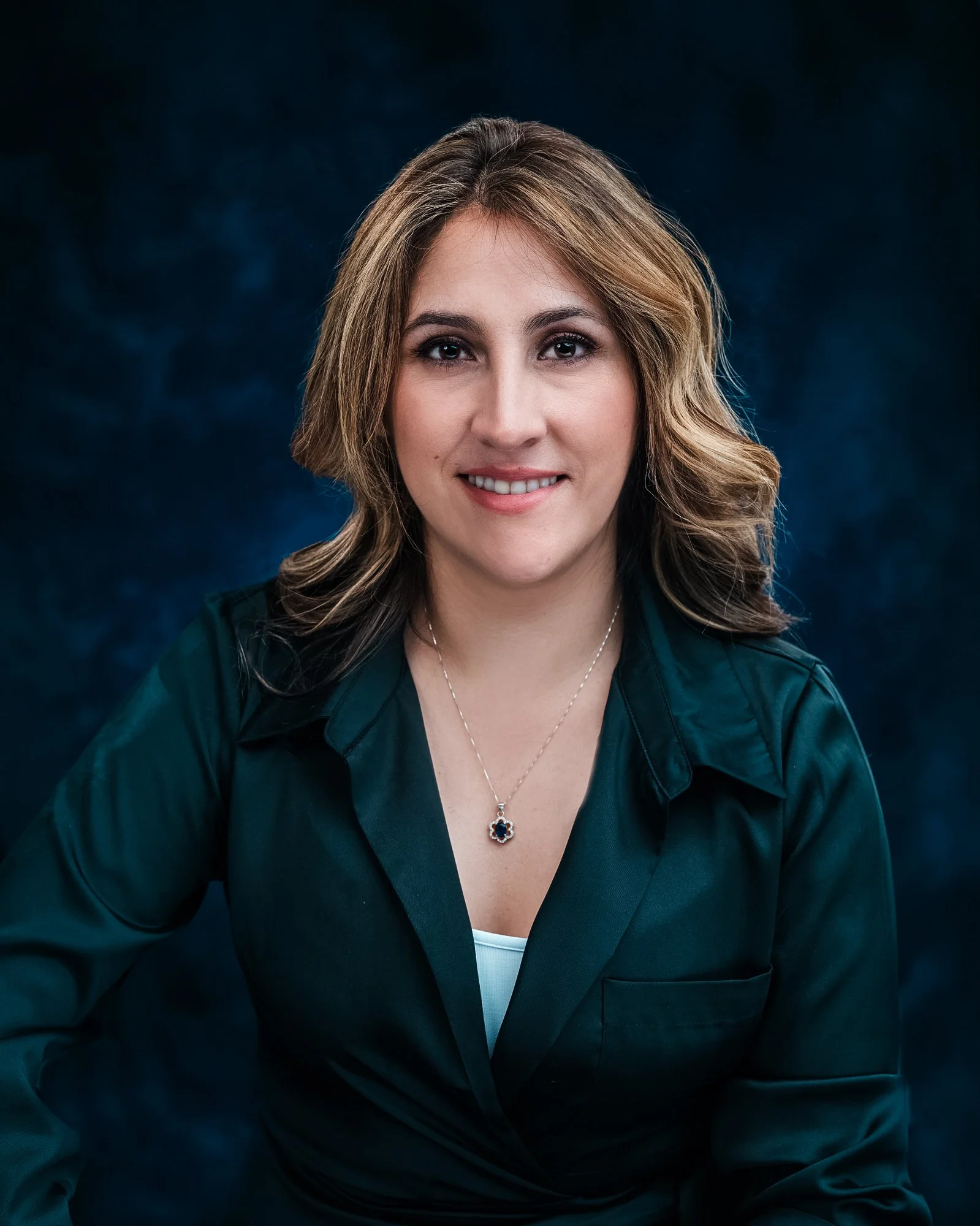 A woman with wavy, shoulder-length brown hair, wearing a dark teal blazer and a silver necklace with a dark blue gemstone pendant, smiling at the camera against a dark blue background.