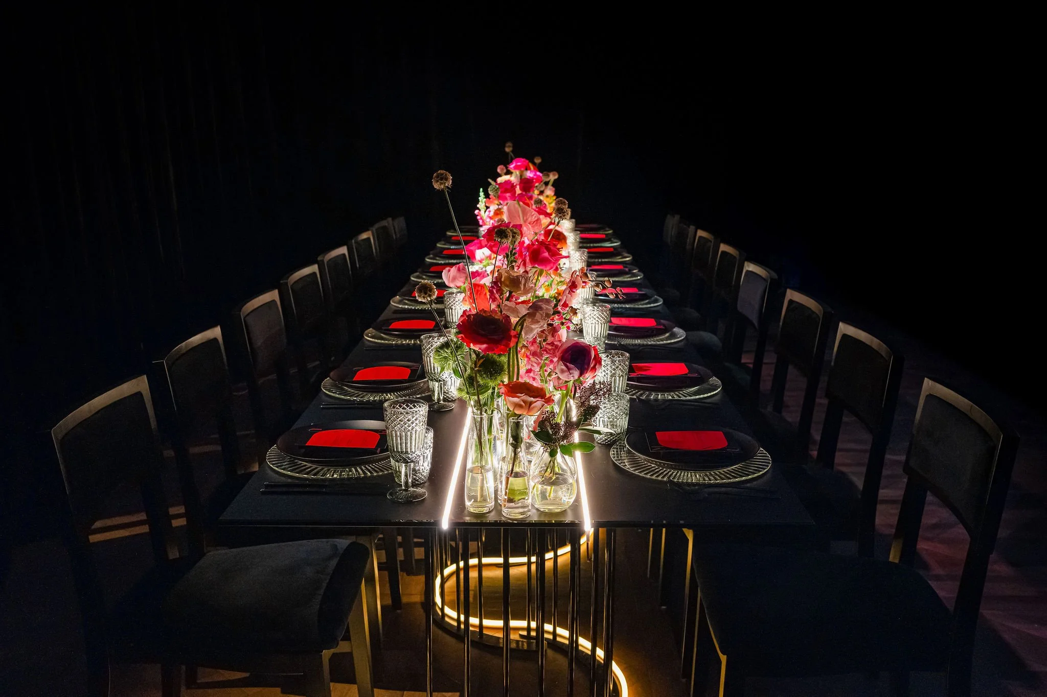A long dining table set for a formal dinner with black tablecloth, pink floral centerpieces, and glassware, illuminated by a gold-lit base.