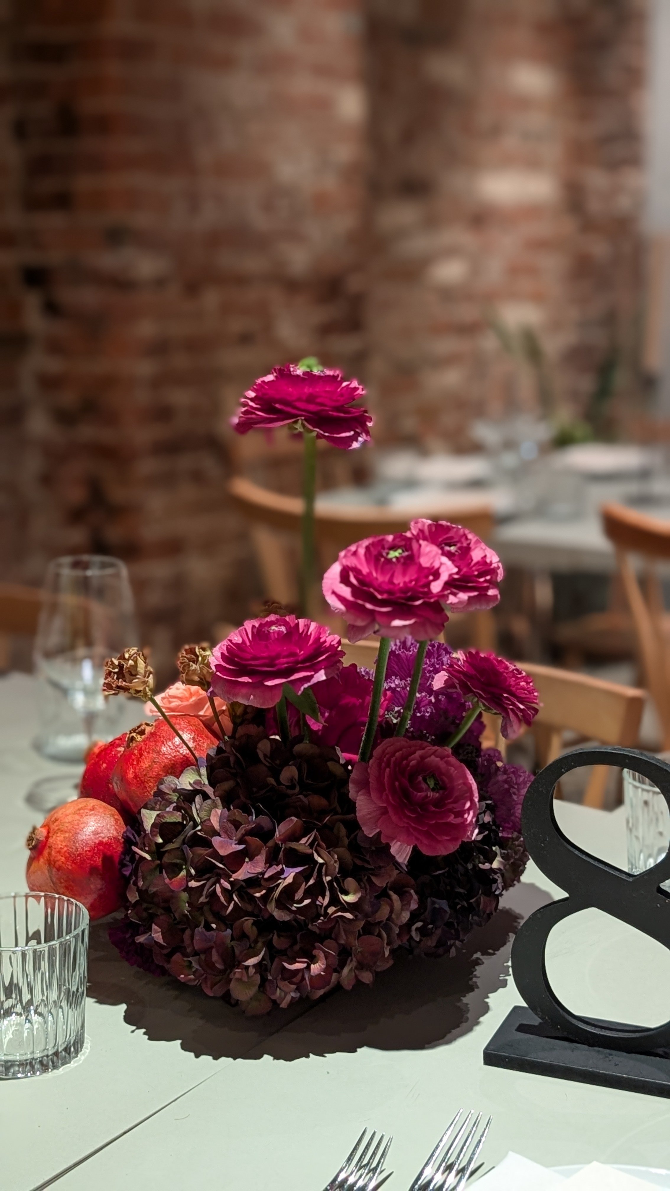 A floral centerpiece with pink and purple flowers, pomegranates, and candles on a white table in a dining setting with a brick wall background.