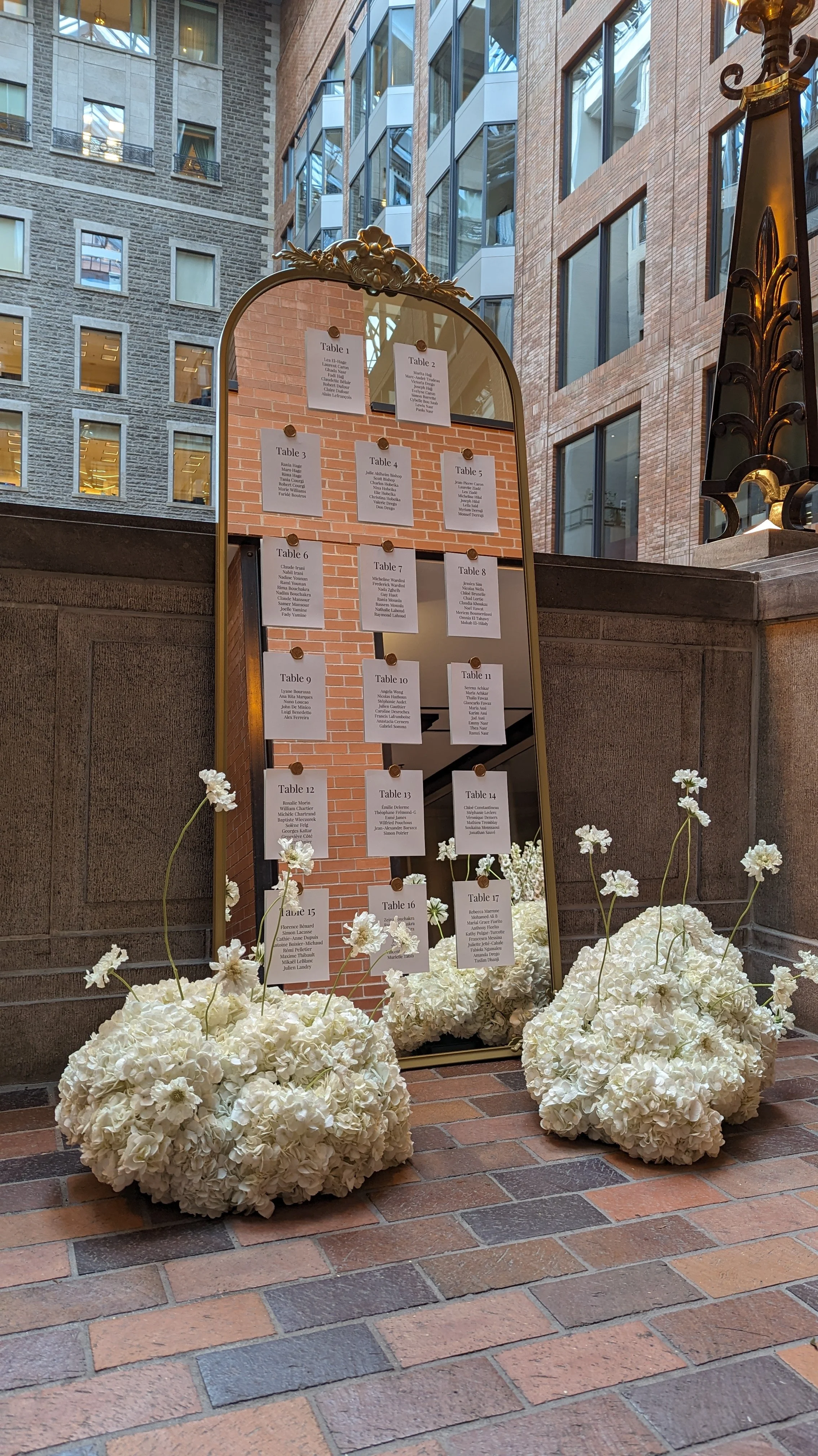A mirror with a golden ornate frame displaying a seating chart for an event, placed on a brick floor in a lobby or atrium with tall glass windows and brick walls, surrounded by white floral arrangements.