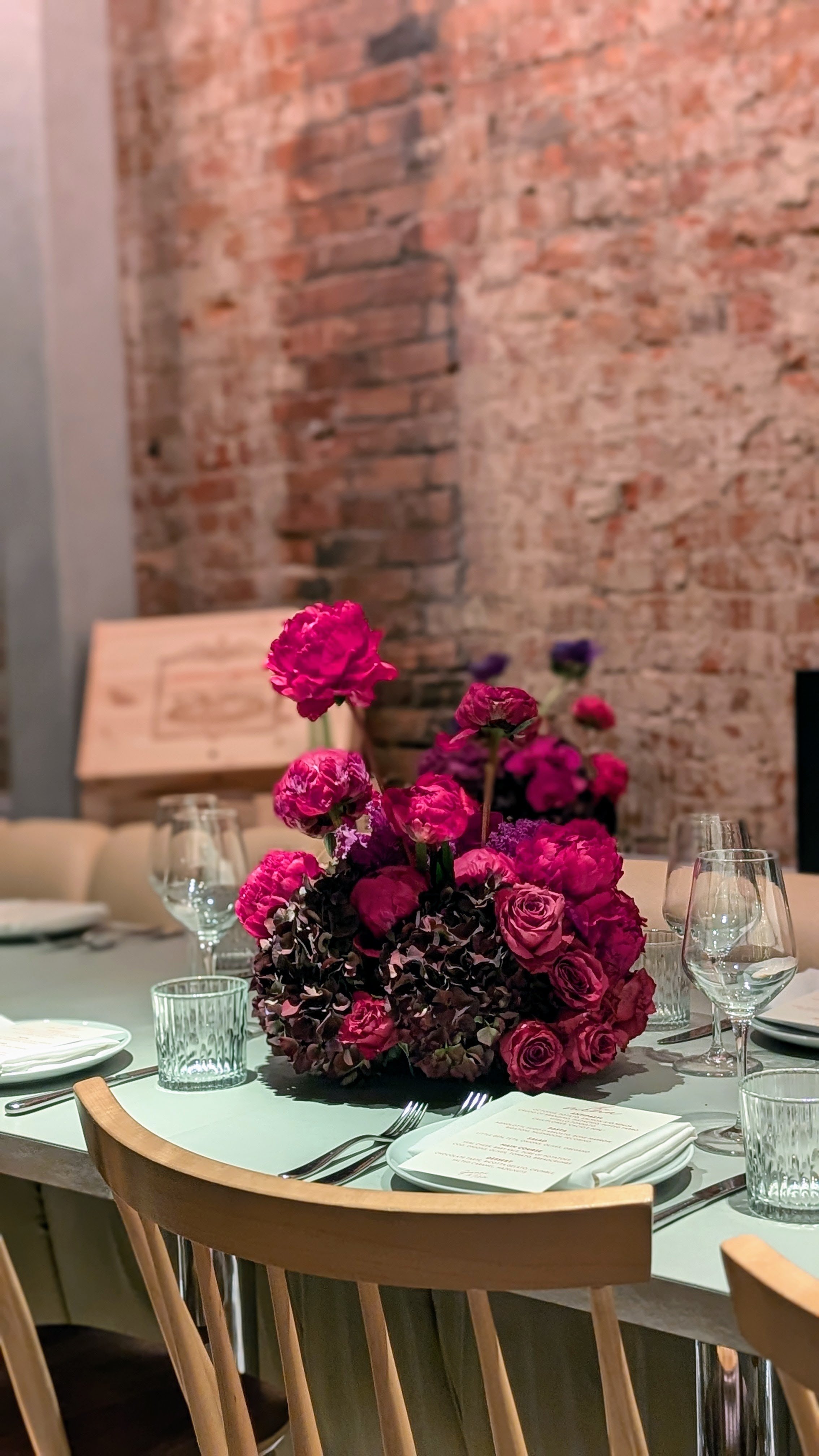 A dining table set for a formal meal with a large pink floral centerpiece, wine glasses, water glasses, plates, cutlery, and menus, in a room with brick walls and wooden chairs.