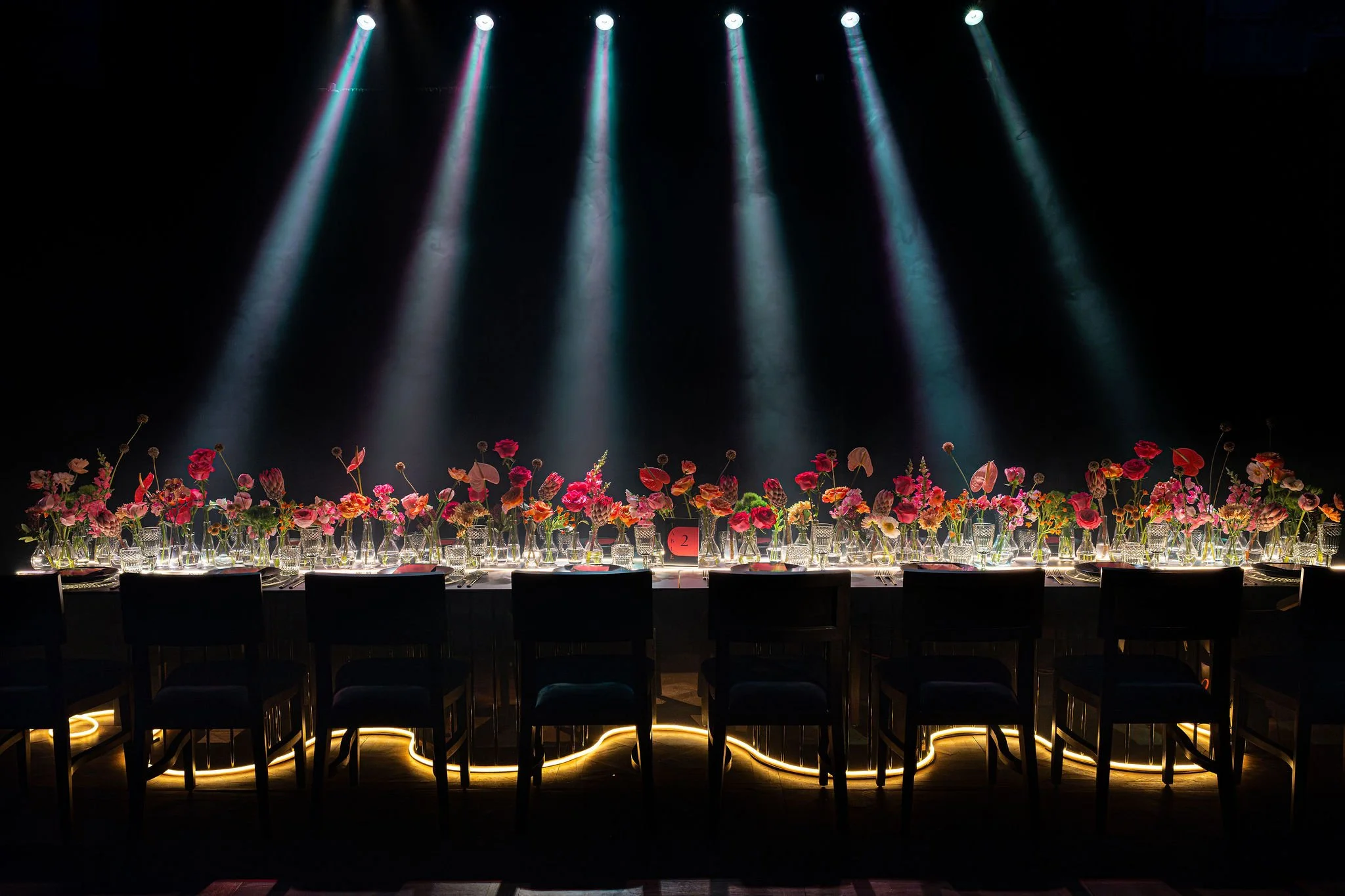 Long table decorated with pink, red, and orange flowers in glass vases, set with glassware and flatware, illuminated by string lights beneath, with spotlights shining down from above in a dark room.
