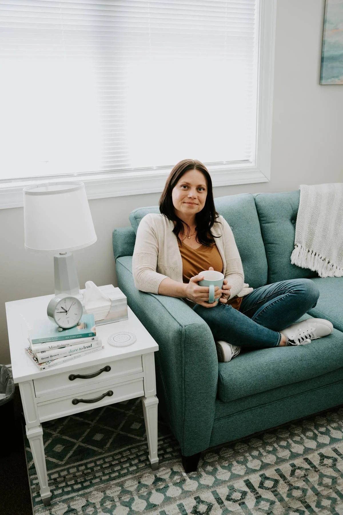 A counselor is sitting cross-legged on a blue couch while holding a coffee mug. Counselor looks warmly at the camera.