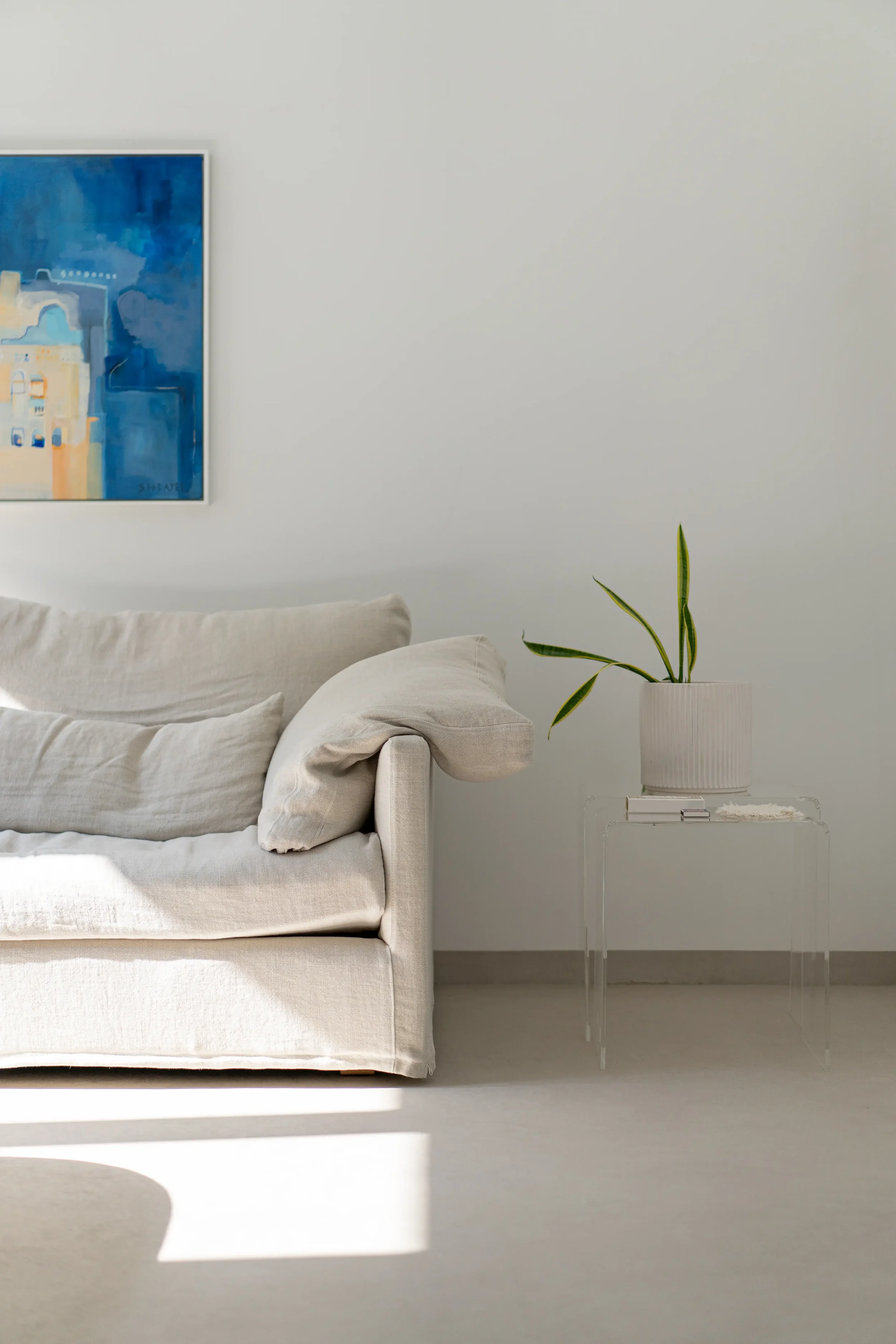 Minimalist living room with a beige sofa, a white side table with a potted green plant, a blue abstract painting on the wall, and natural sunlight streaming in.