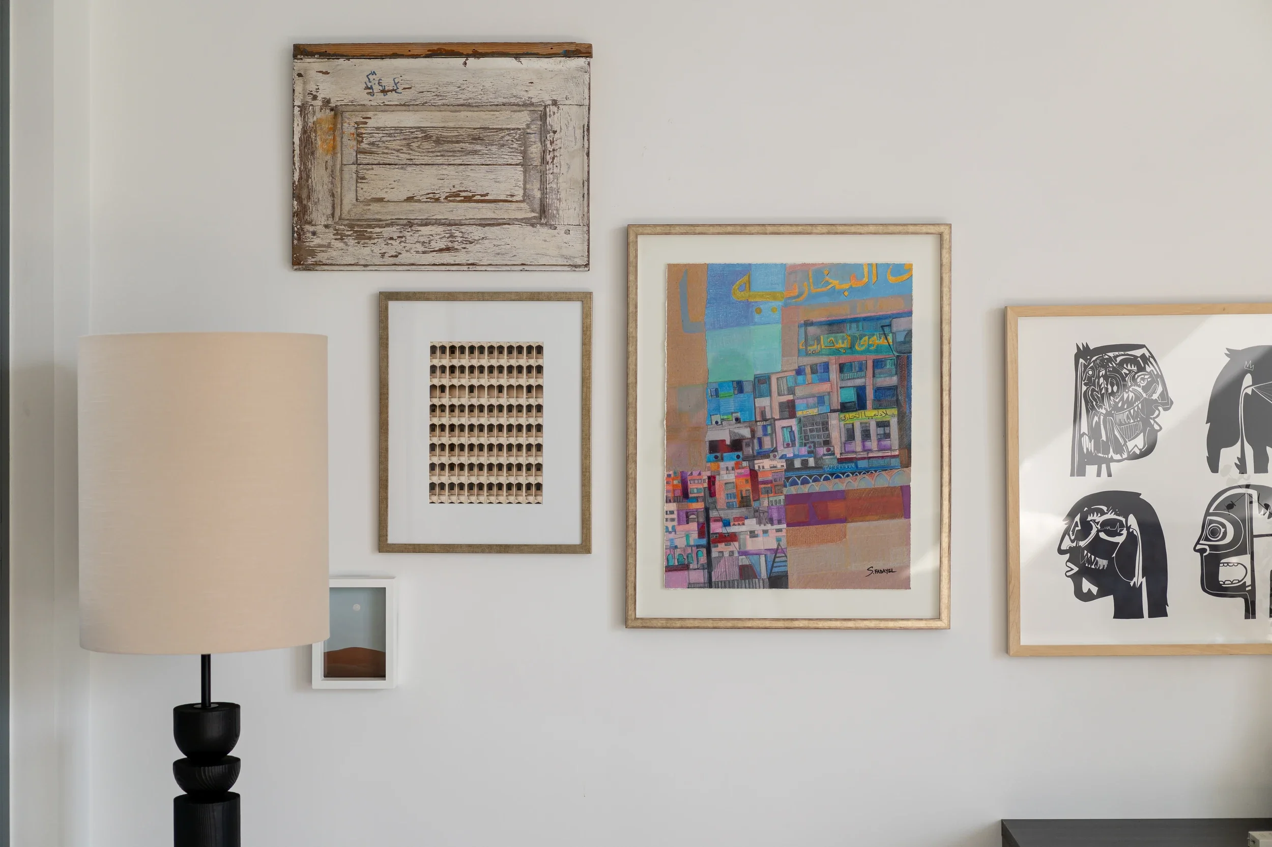 A wall with various framed artworks, including a rustic wooden frame, a grid of small objects, a colorful abstract painting with Arabic script, and a piece with black and white profiles of animals or faces, with a beige lamp on a black stand on the left.