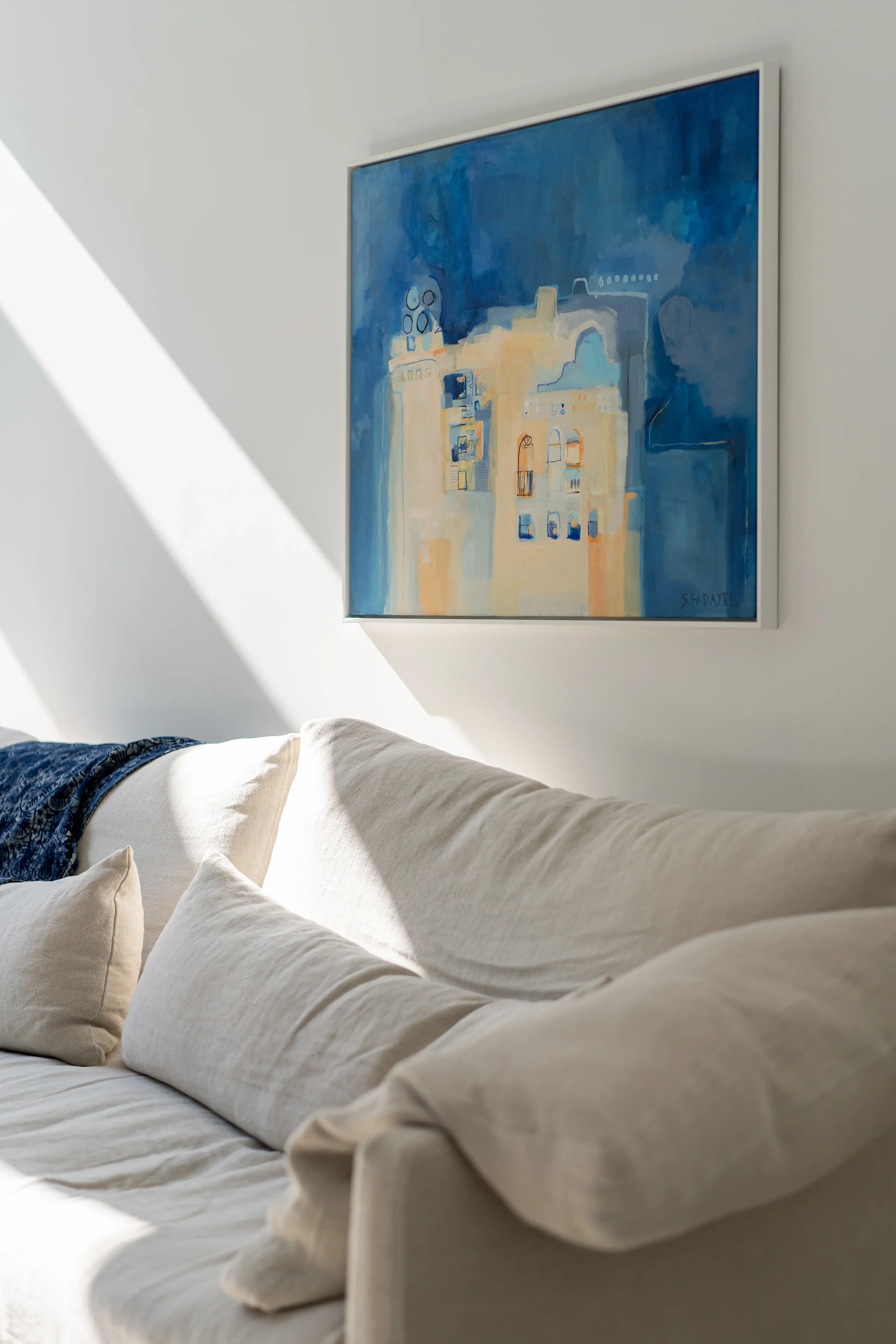 A beige sofa with cushions in front of a white wall with a framed abstract painting in shades of blue, yellow, and white, sunlight casting shadows.