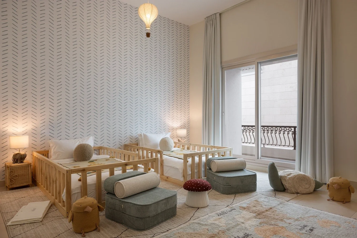 A children's bedroom with two small beds made of light wood, each with white pillows and soft spherical cushions. The room has a patterned accent wall, a large sliding glass door with white curtains leading to a balcony, and children's toys and seating on the floor, including ottomans, a mushroom-shaped stool, plush animal-shaped baskets, and a soft rug.
