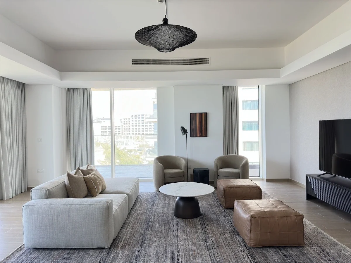 Modern living room with white sofa, two beige armchairs, a round coffee table, two brown leather ottomans, a flat-screen TV, and large windows with light-colored curtains, overlooking city buildings.