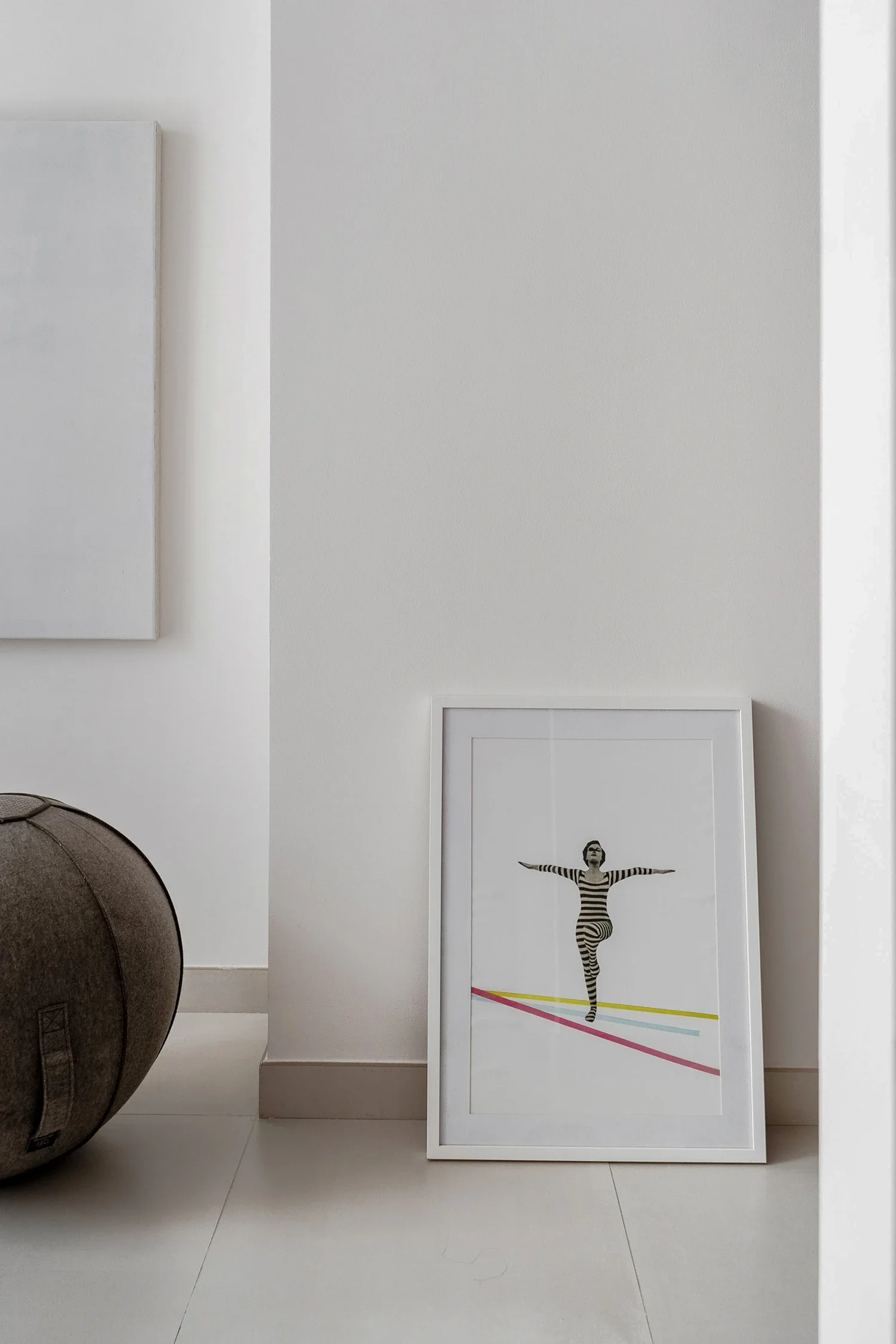 Framed black and white photo of a person in striped clothing performing a yoga pose, leaning on one leg, inside a white frame, placed on the floor next to a gray spherical bag, against a white wall.