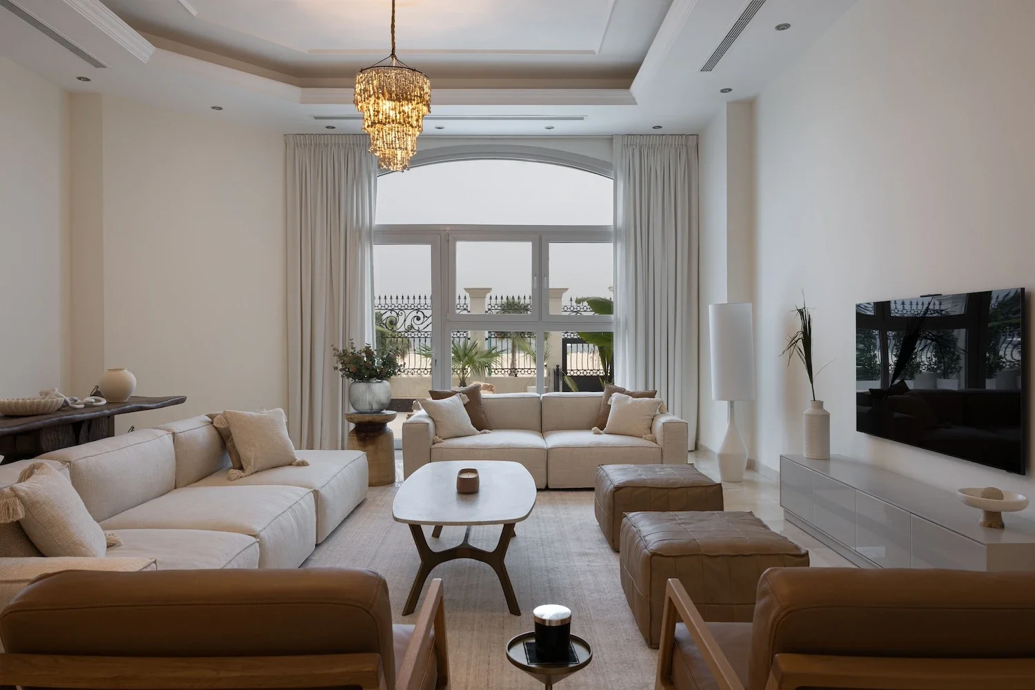 A modern living room with white couches, a wooden coffee table, a large window with white curtains, and a flat-screen TV mounted on the wall.