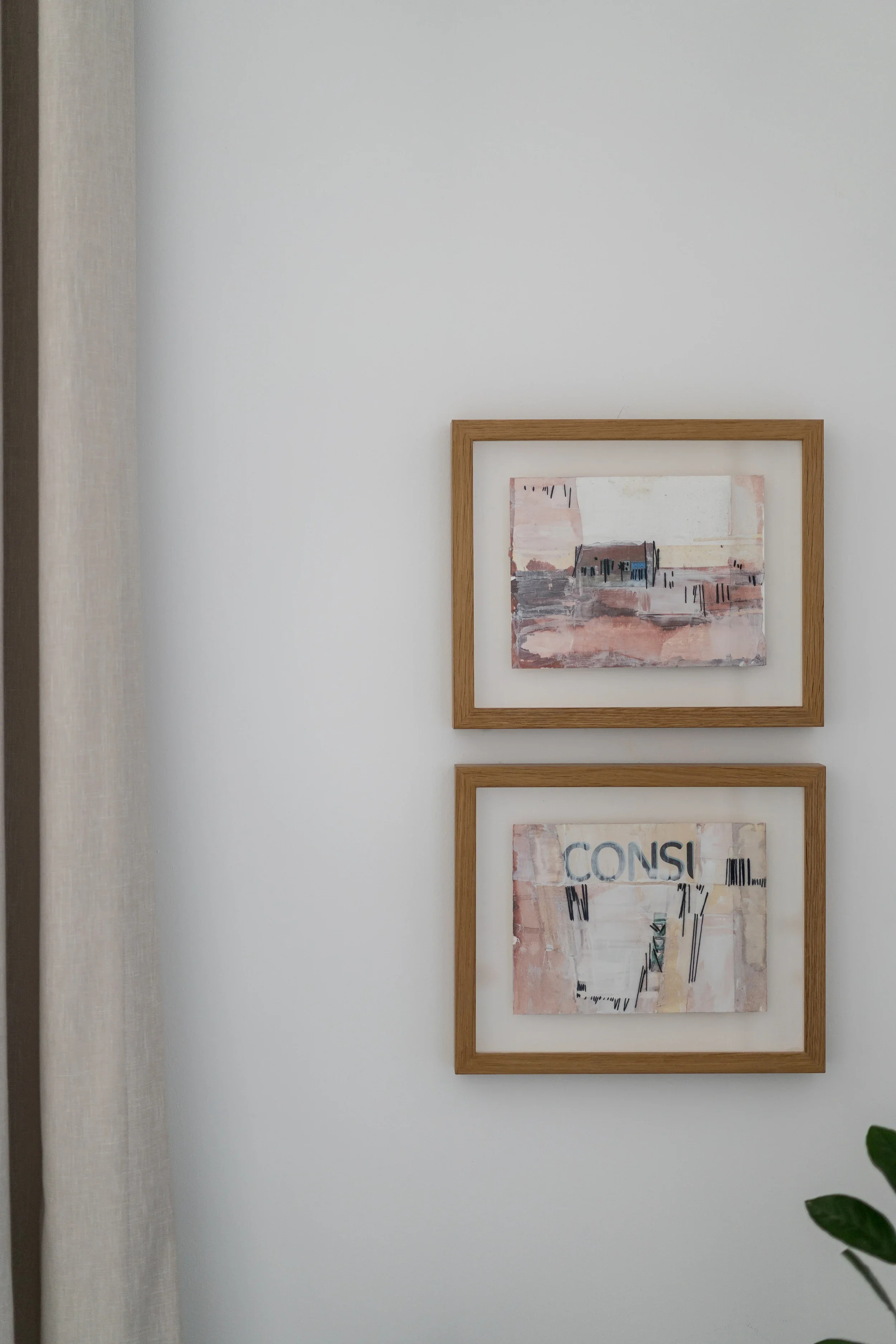 Two framed abstract paintings on a white wall with a beige curtain partially visible on the left and a green plant in the lower right corner.