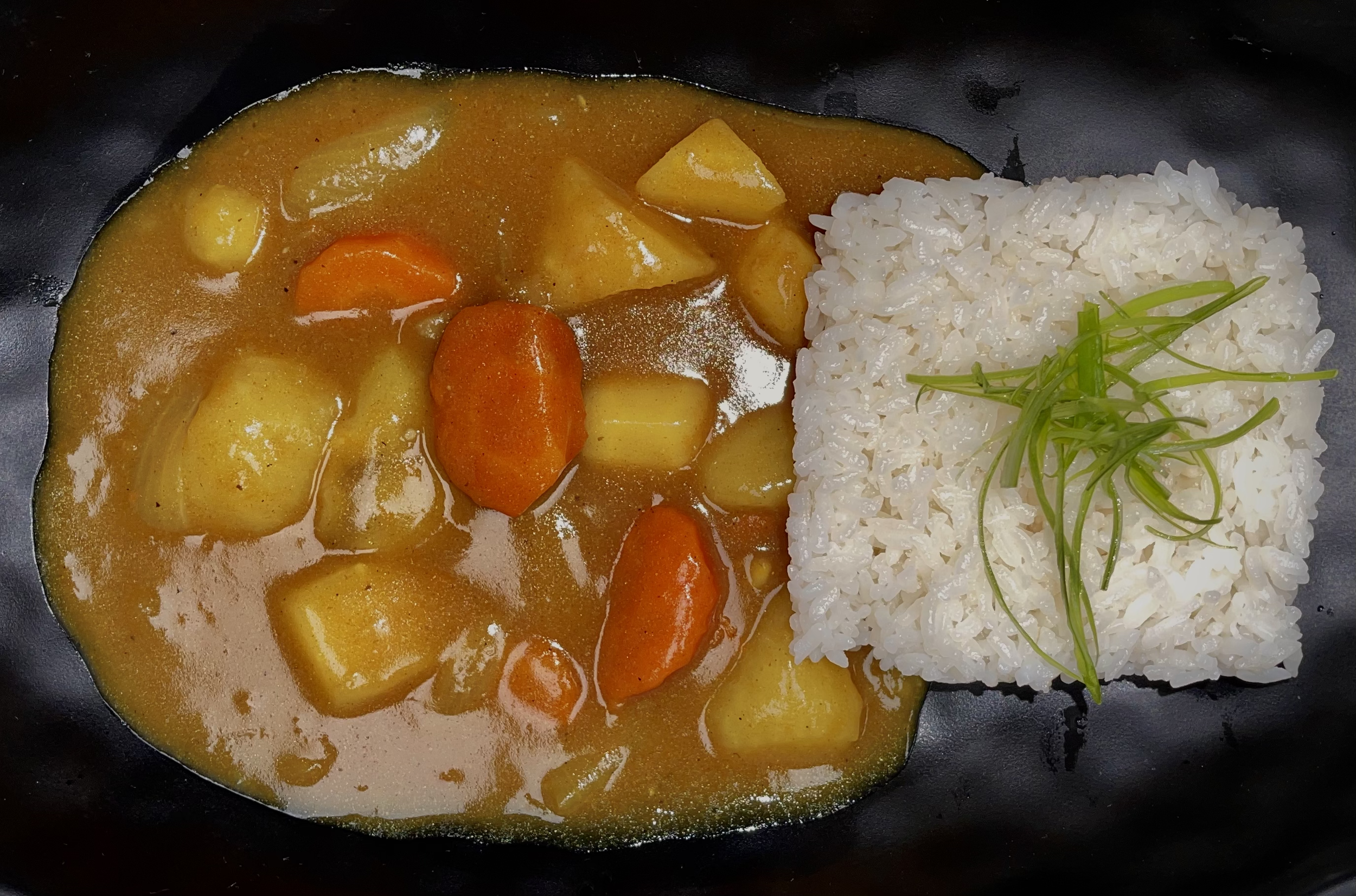 A black plate with serving of Japanese curry containing potatoes and carrots, alongside a square portion of steamed white rice garnished with green herbs.