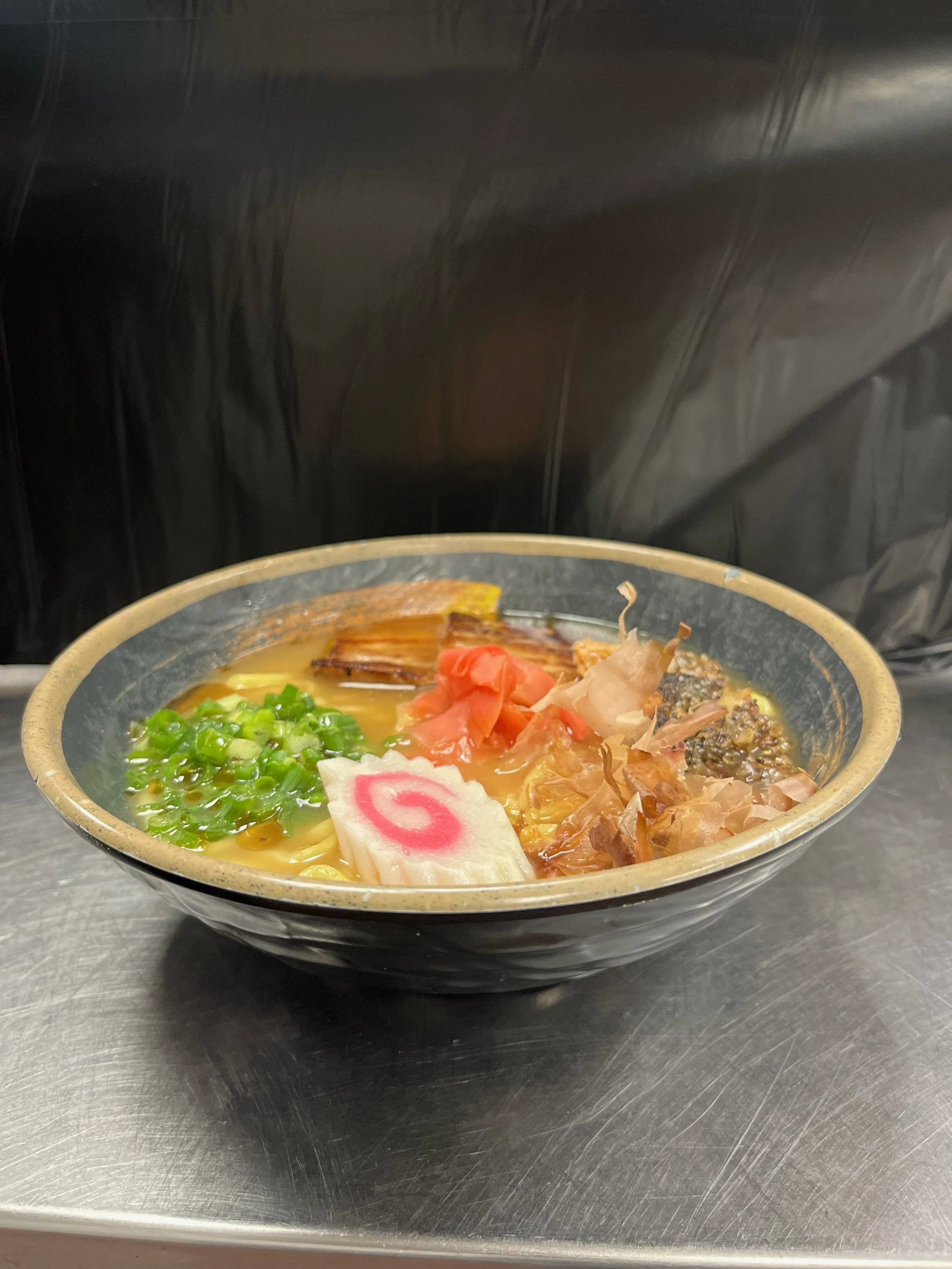A bowl of ramen noodles topped with green onions, pink narutomaki, slices of pork, pickled ginger, seaweed, and bonito flakes.