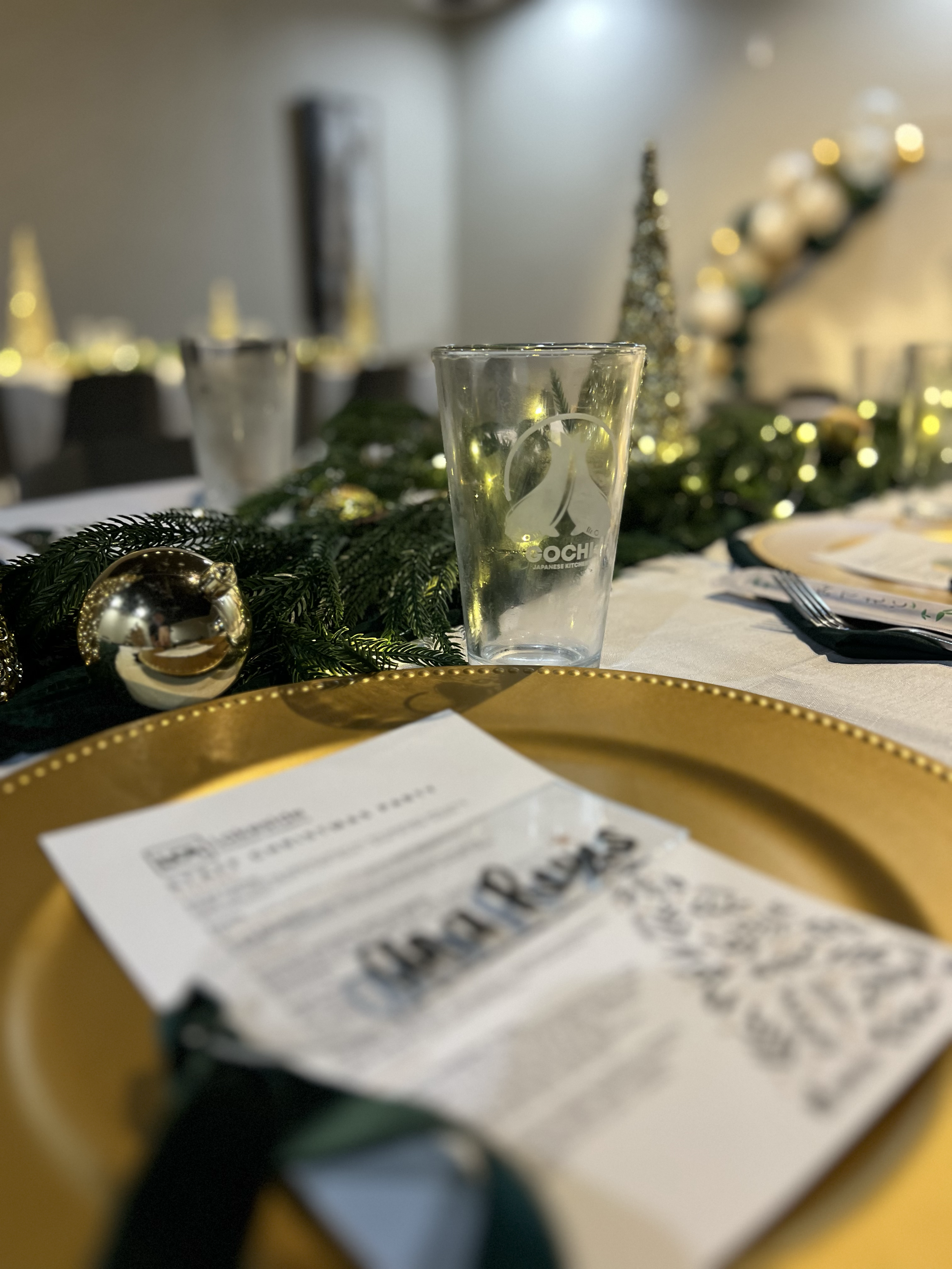 A festive table setting with a gold charger plate, a menu with a metal clip, a clear glass, and holiday decorations including a pine garland and gold ornaments, in a room decorated for Christmas.