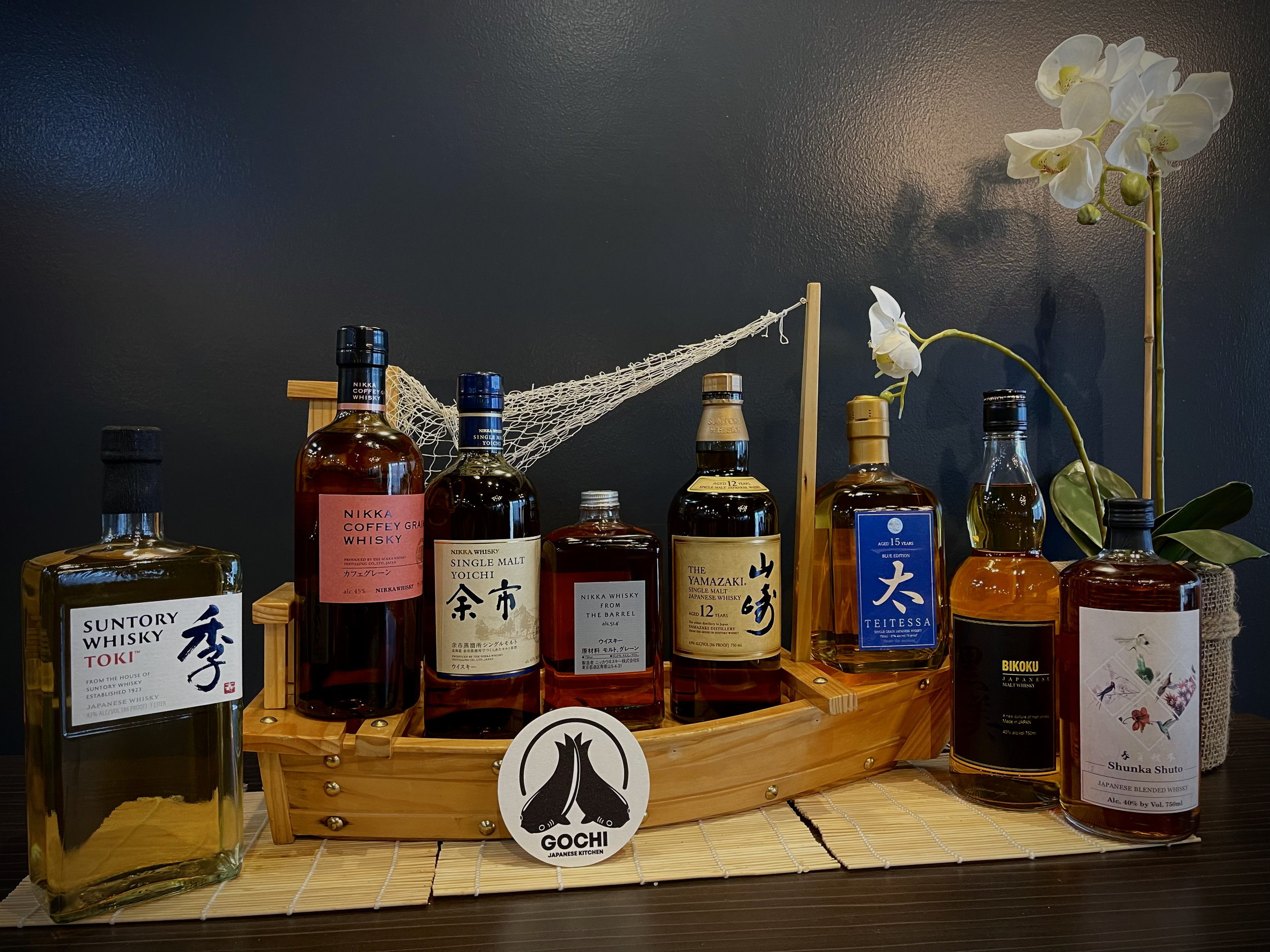 Assorted bottles of Japanese whiskey displayed on a wooden tray, with orchids and a small decorative sign that says 'GOCHI' in front.