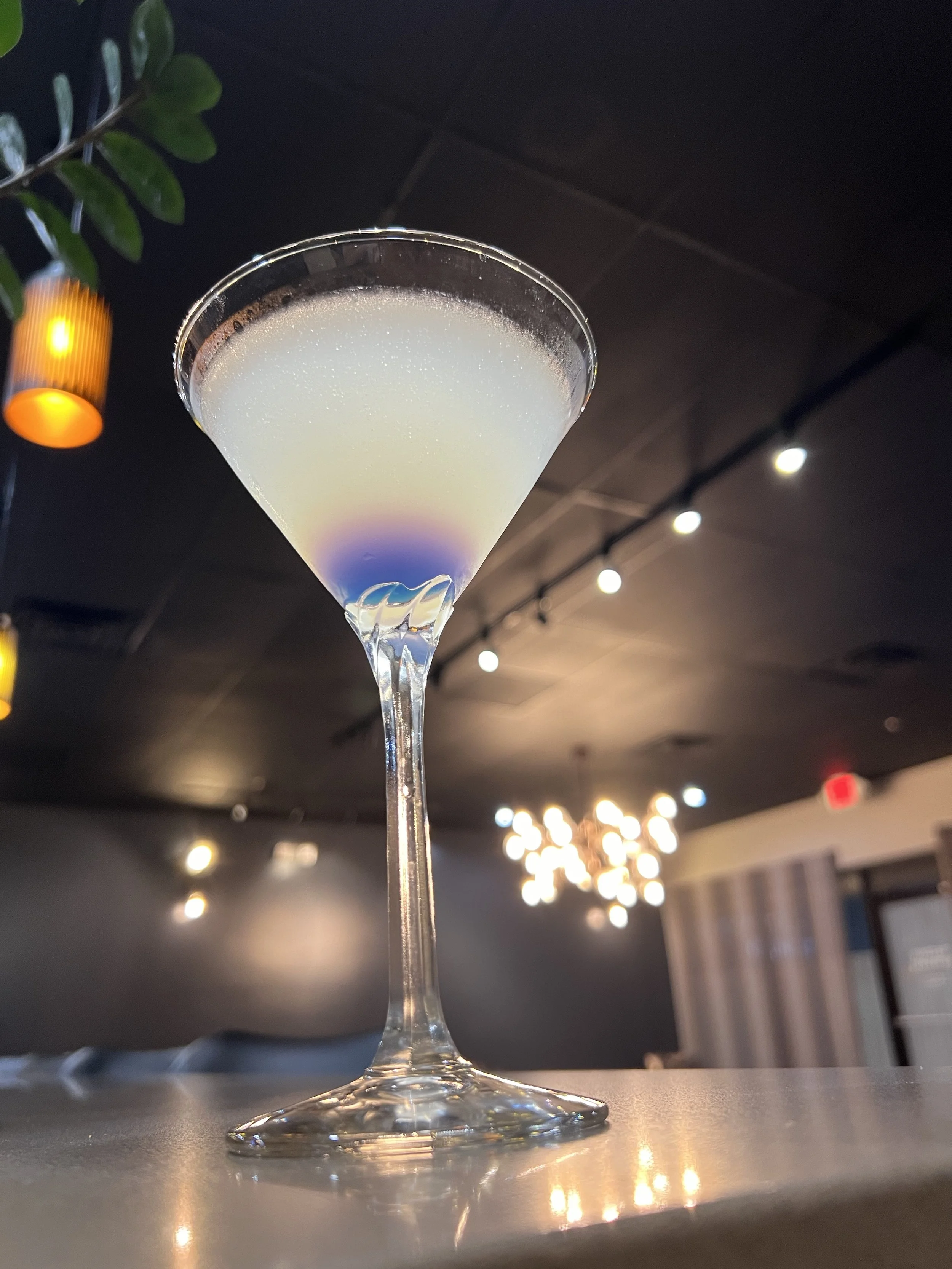 A cocktail glass filled with a light purple or lavender-colored drink, placed on a bar or counter in a dimly lit restaurant or bar setting with modern decor and hanging lights in the background.
