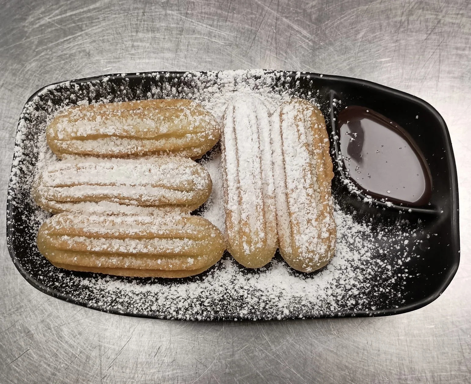 A black tray with three ladyfinger cookies dusted with powdered sugar, a small bowl of chocolate sauce, and three more ladyfingers dusted with powdered sugar on a silver surface.