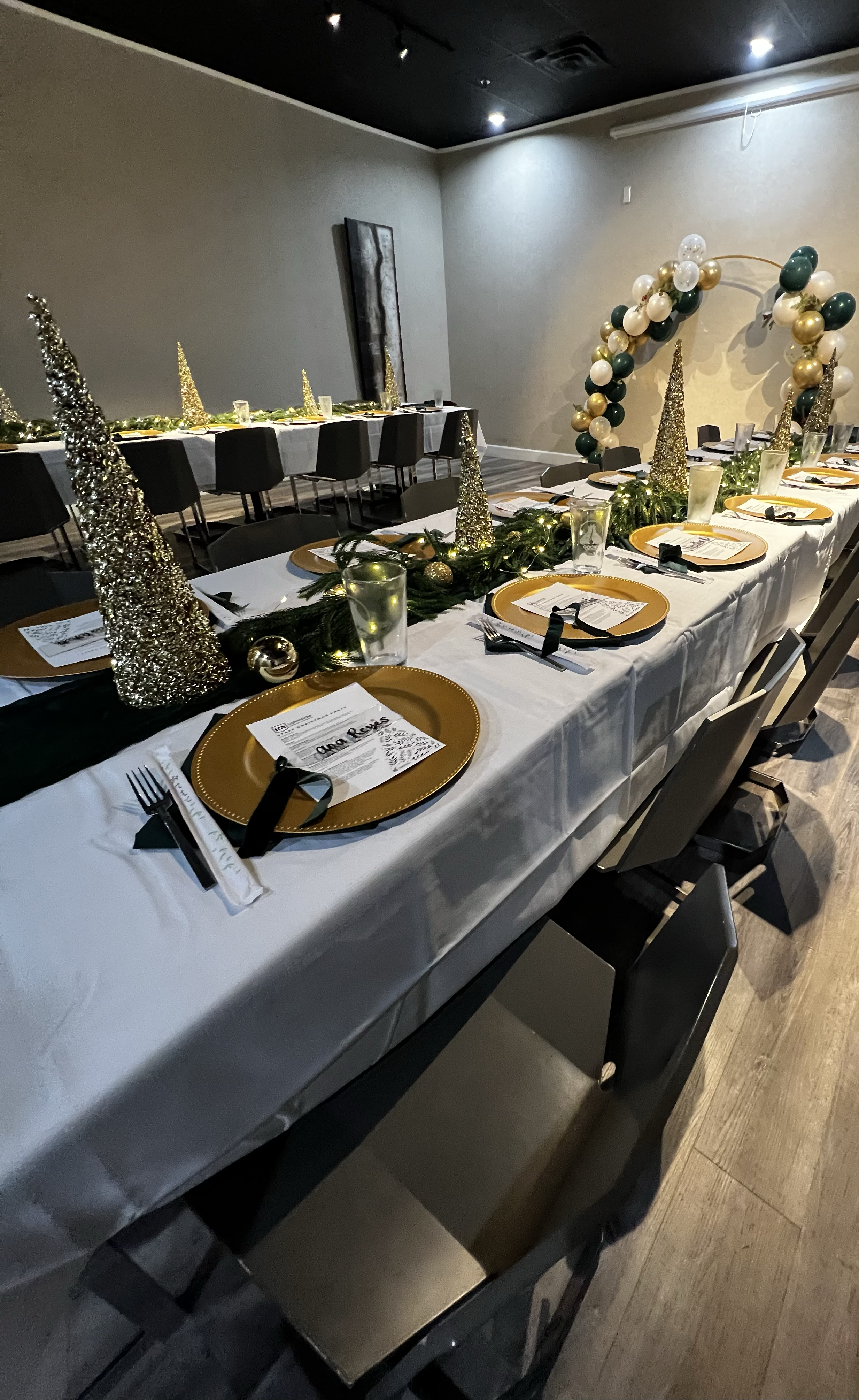 A long rectangular table decorated for a holiday celebration with gold and silver cone-shaped ornaments, greenery, and string lights as a centerpiece. The table is set with gold chargers, printed menus, glasses, and utensils, and has black and white 