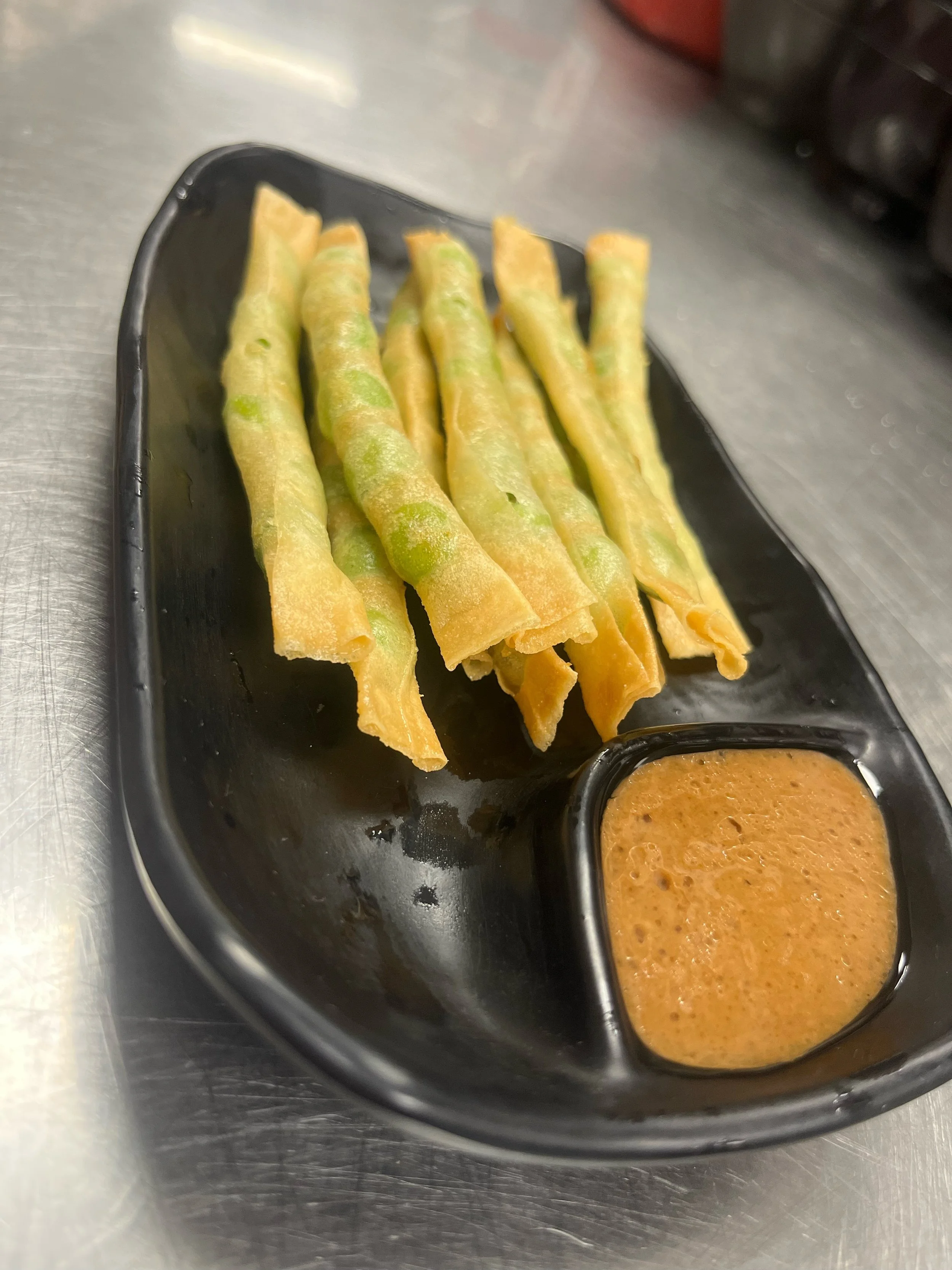 Fried wonton wrapped edamame sticks served on a black plate with a side of dipping sauce.