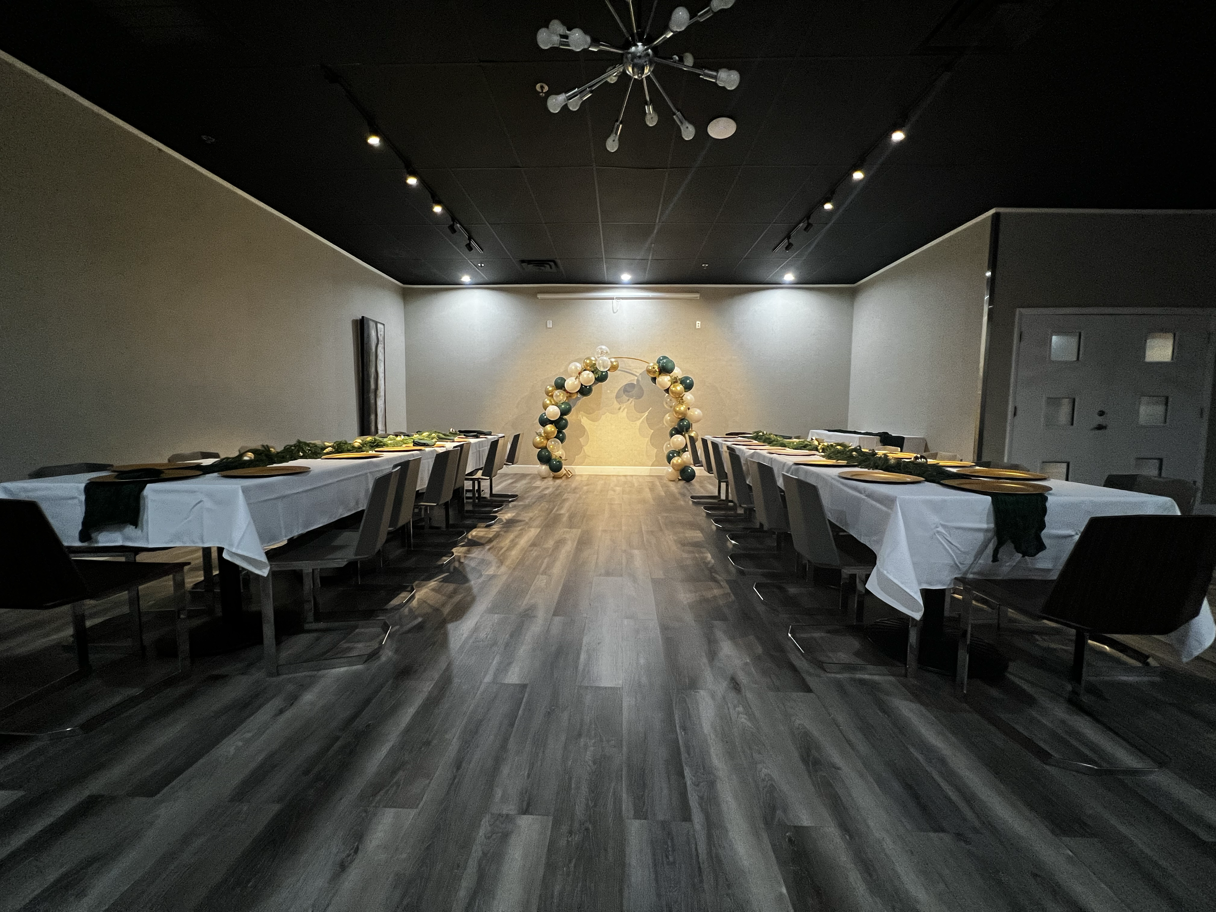 A decorated event space with two long banquet tables covered with white tablecloths and black napkins, set with plates and cutlery. The tables are adorned with green garlands. At the back of the room, there is a balloon arch made of gold, black, and 