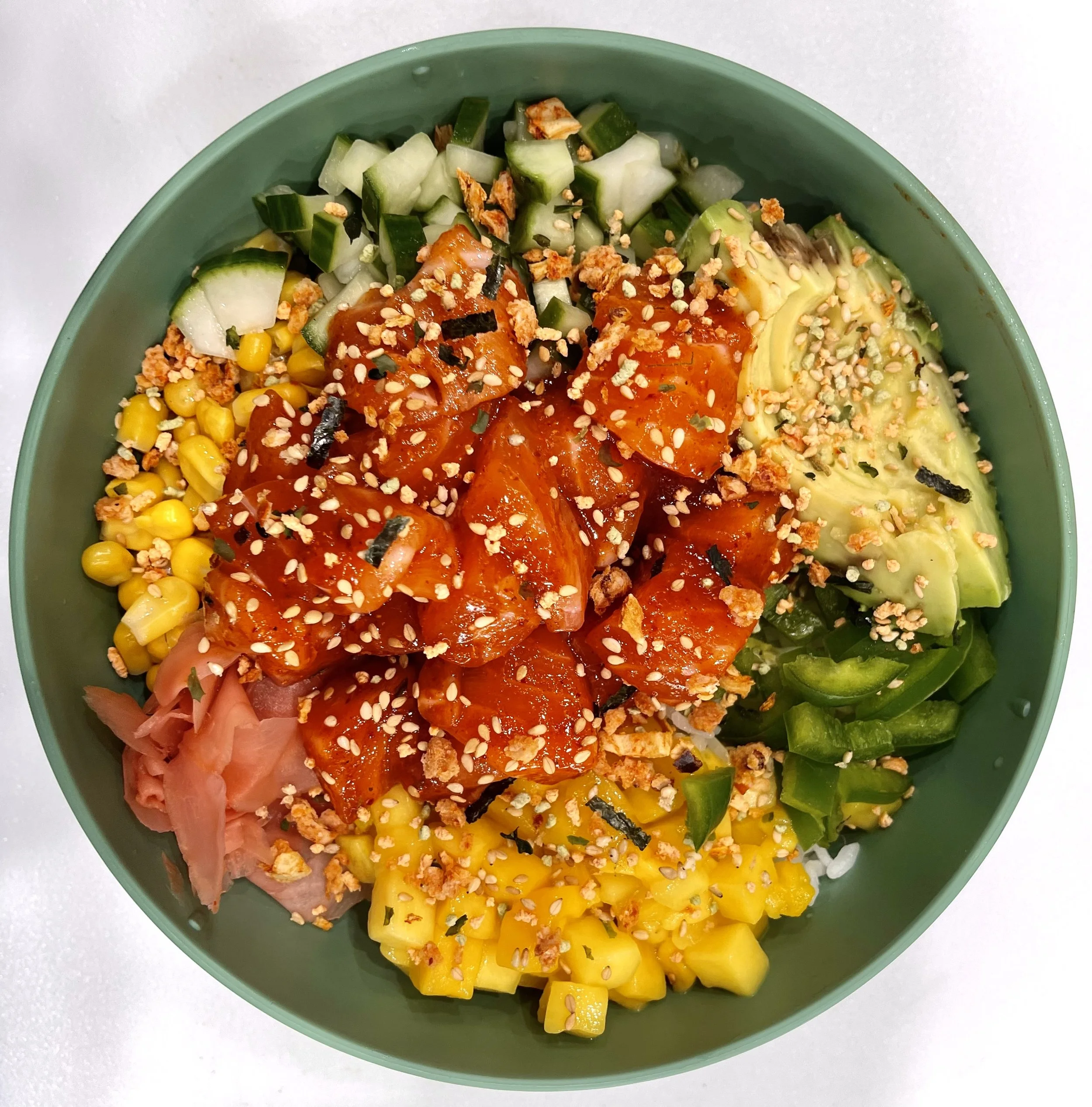 A bowl filled with a colorful poke salad containing diced cucumbers, corn kernels, diced tomatoes, sliced avocado, chopped green bell peppers, pickled ginger, and sprinkled sesame seeds and crushed nuts.