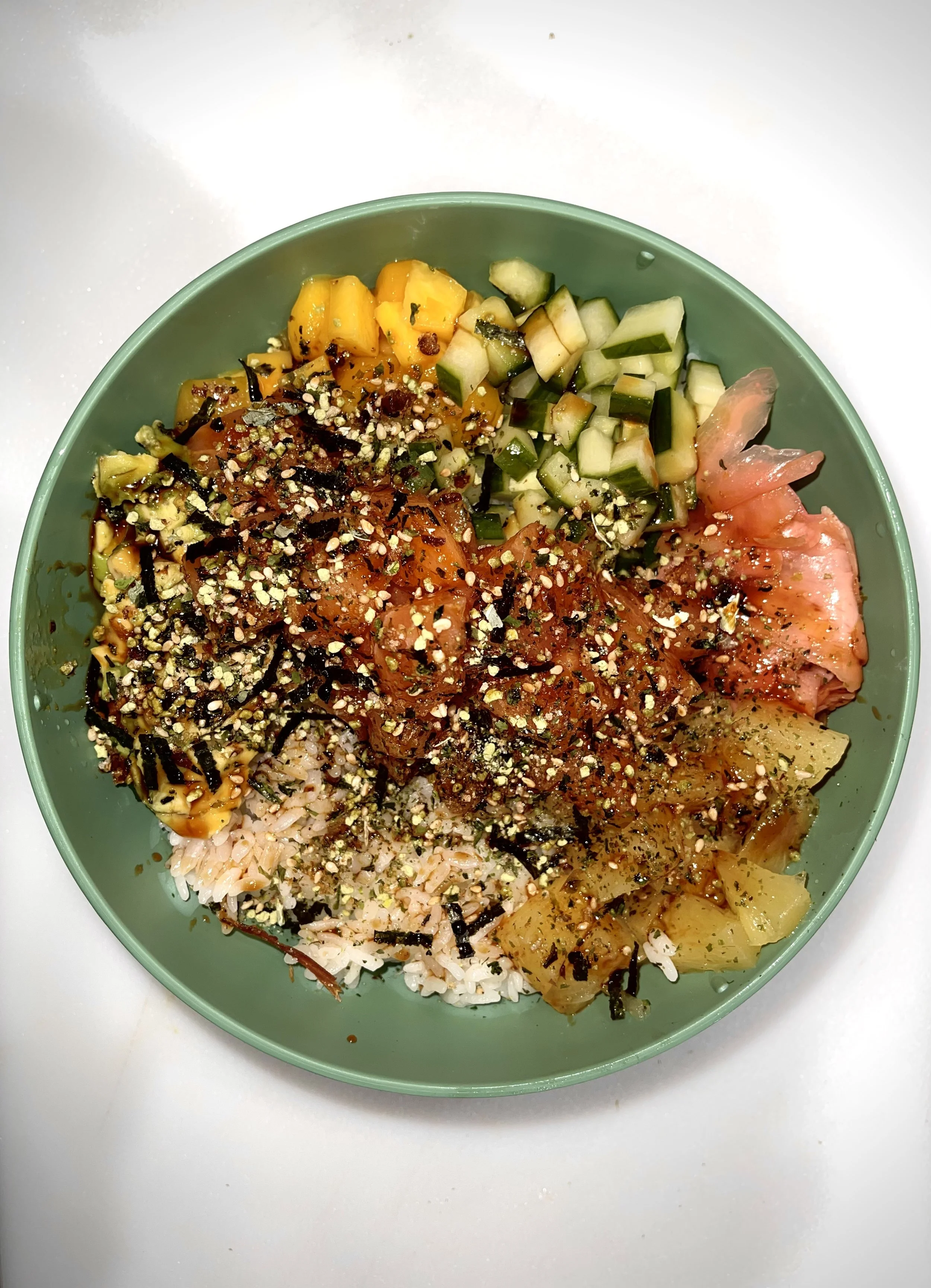 A poke bowl with rice, chopped cucumber, mango, pickled pineapple, ginger, and various toppings including seaweed, crushed nuts, and spicy sauce.