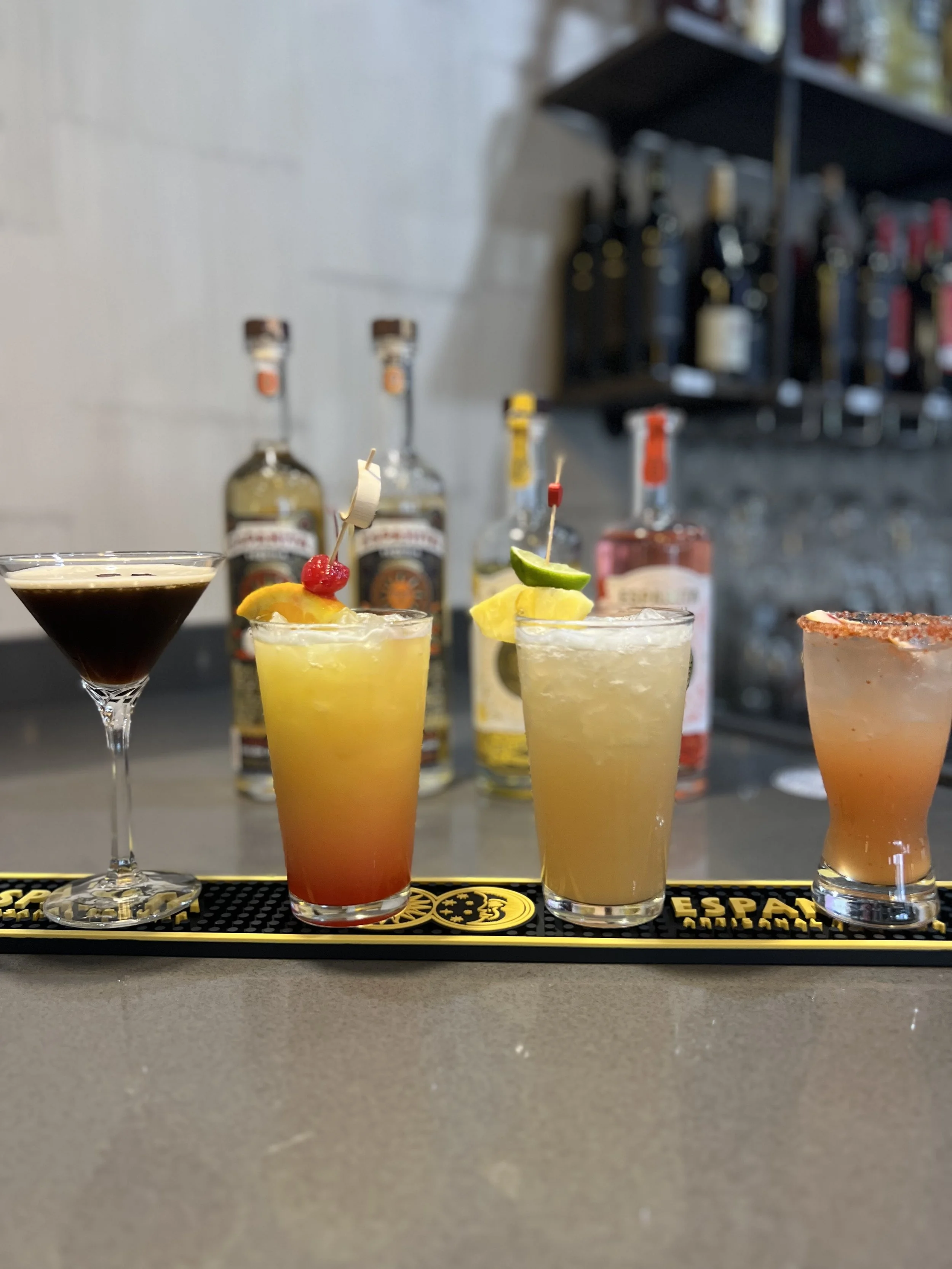 Various colorful cocktails on a bar counter with bottles of alcohol in the background.
