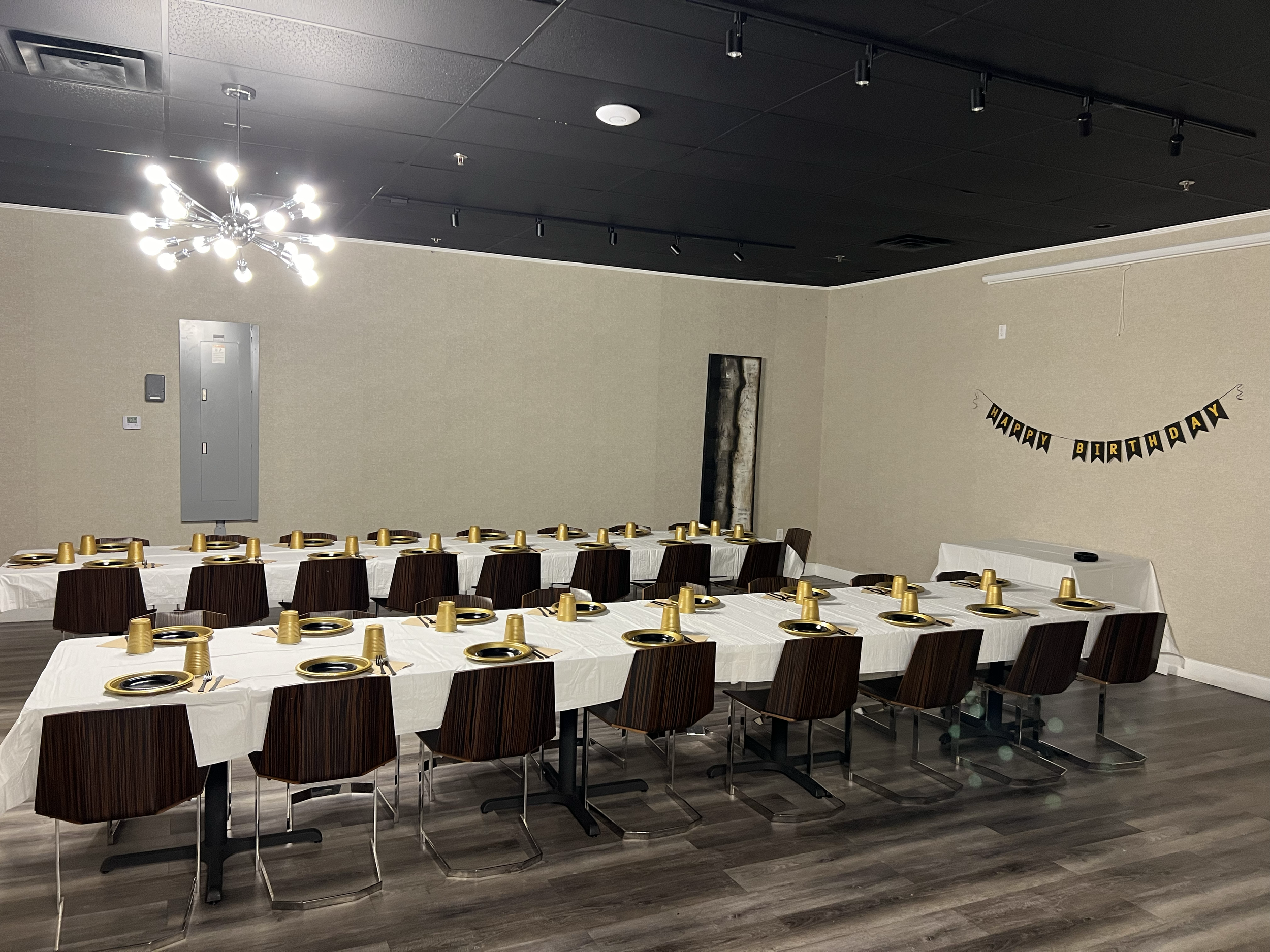 Tables covered with white tablecloths set for a birthday celebration, with gold cups upside down on plates, and a 'Happy Birthday' banner on the wall.
