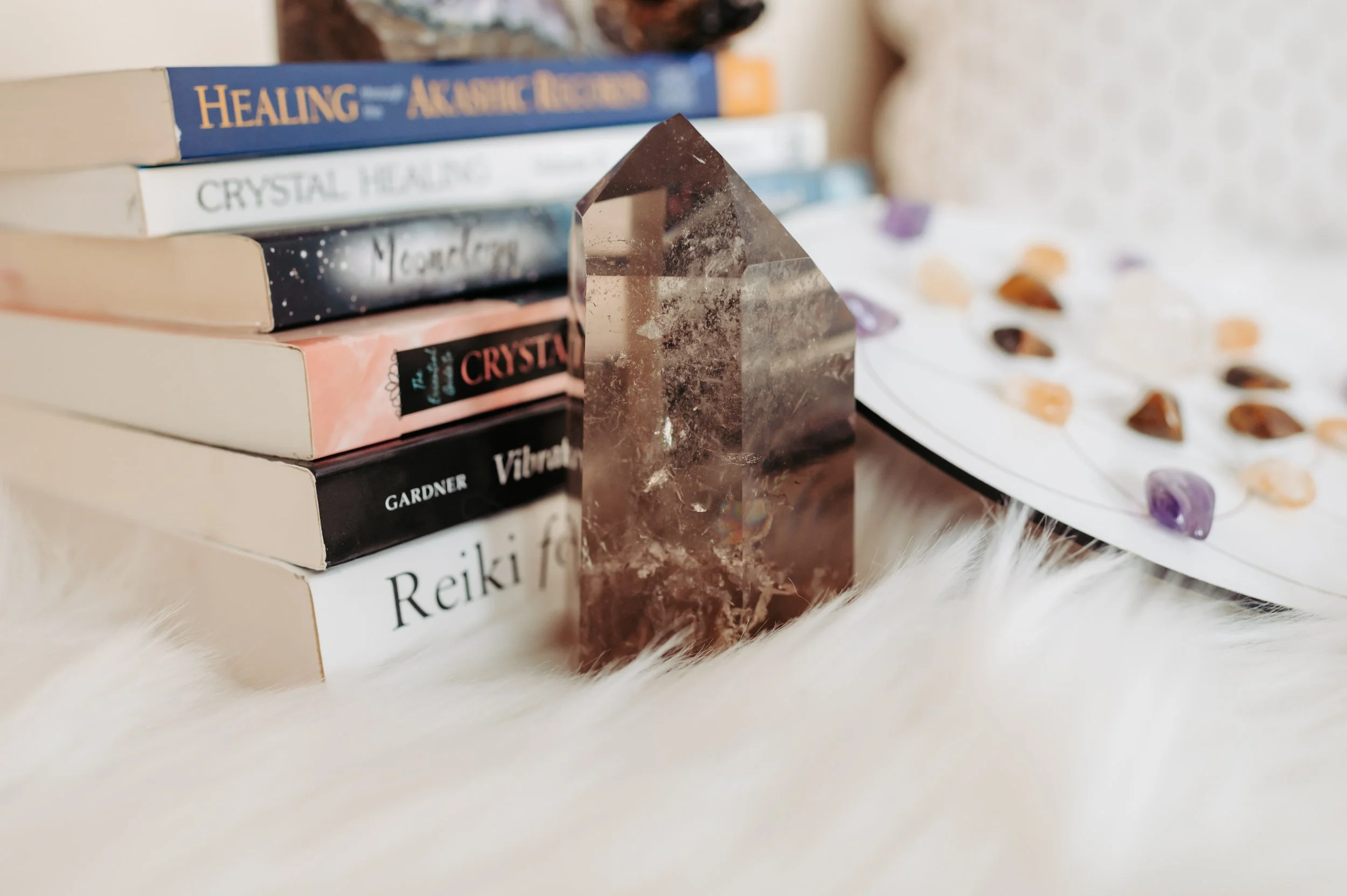 Stack of books on crystal healing and spirituality with a large crystal and chakra stones on a plate, arranged on a fluffy surface.