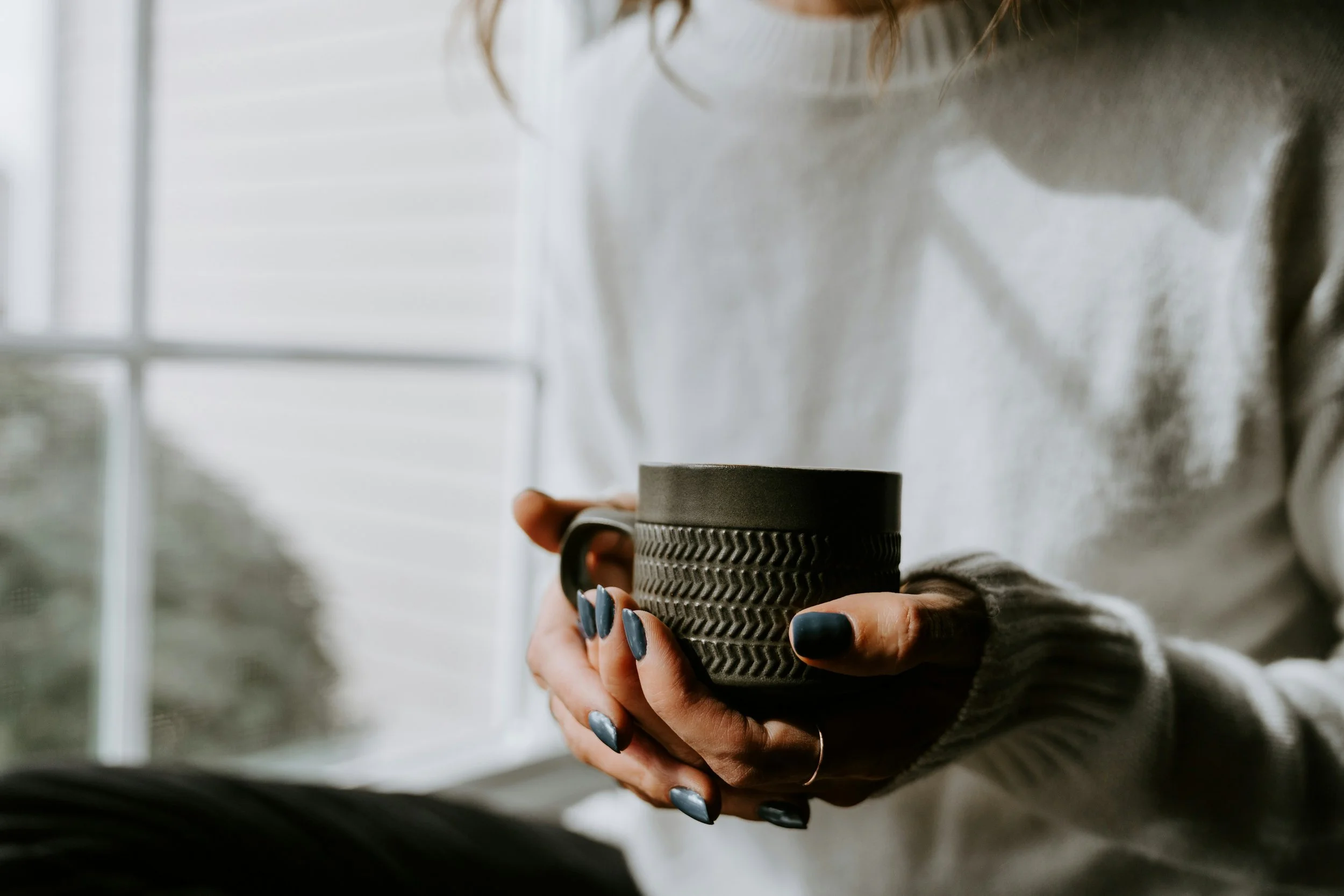 Person holding a textured black ceramic mug, with a background of a window and greenery.