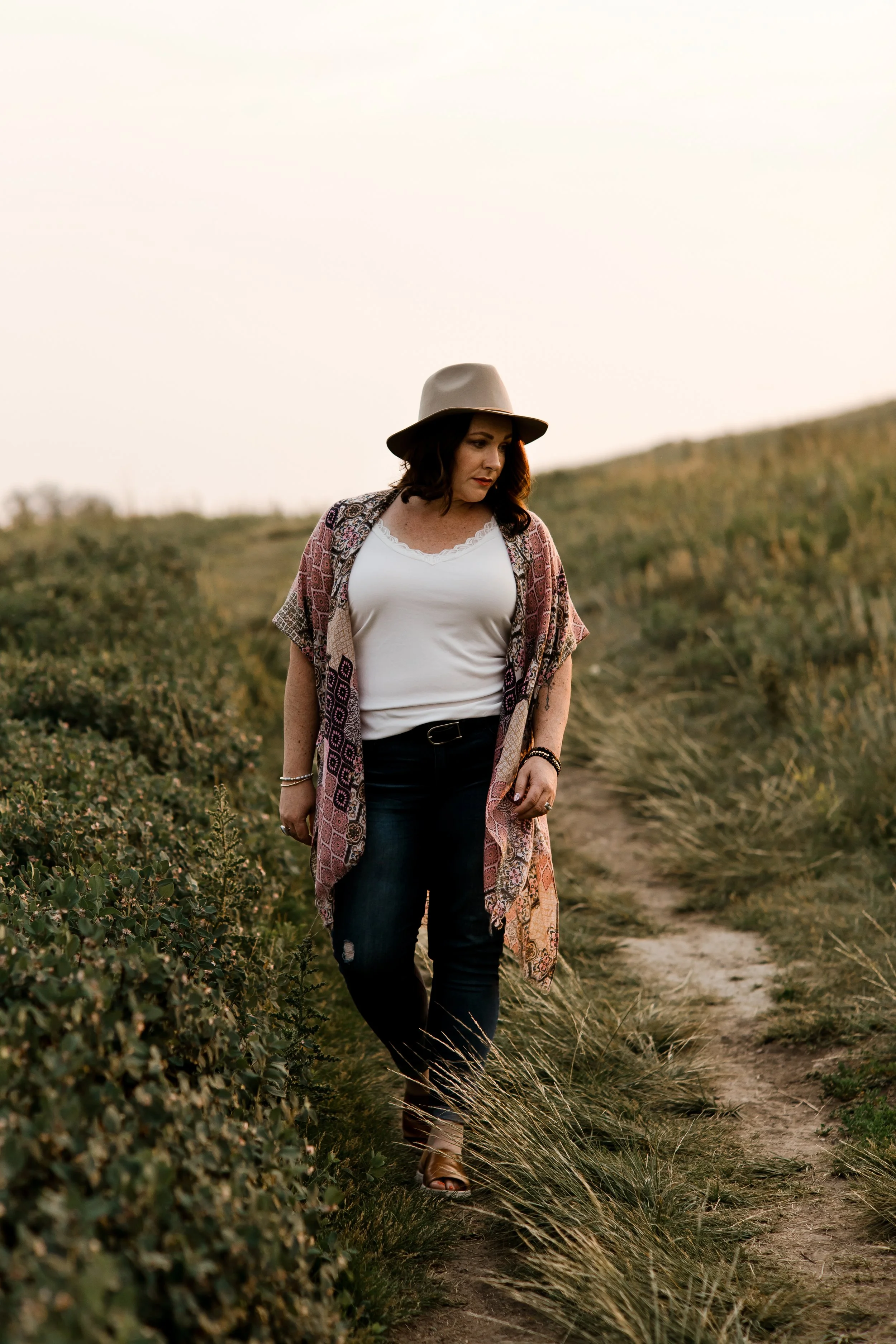 A woman walking on a dirt path through grassy countryside during sunset, wearing a beige hat, white top, patterned kimono, and dark jeans.