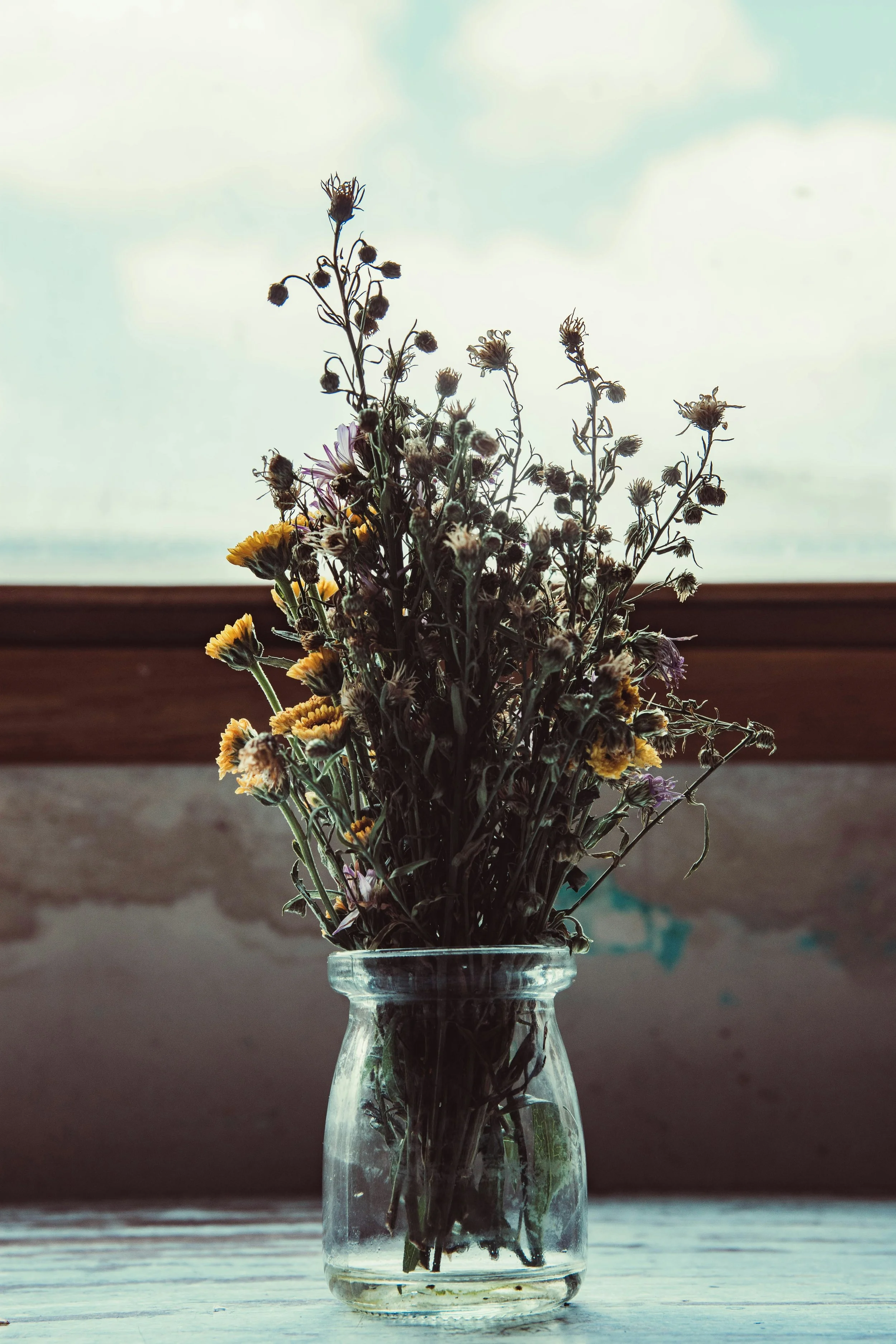 A clear glass jar with a bouquet of dried wildflowers, including yellow, purple, and brown flowers, on a wooden surface with a bright sky background.