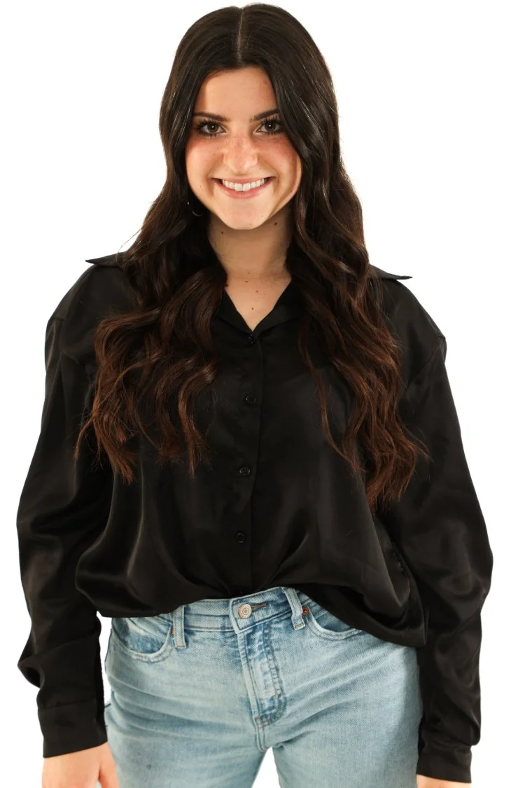 A young woman with long, wavy dark hair smiling at the camera, wearing a black button-up shirt and light blue jeans.