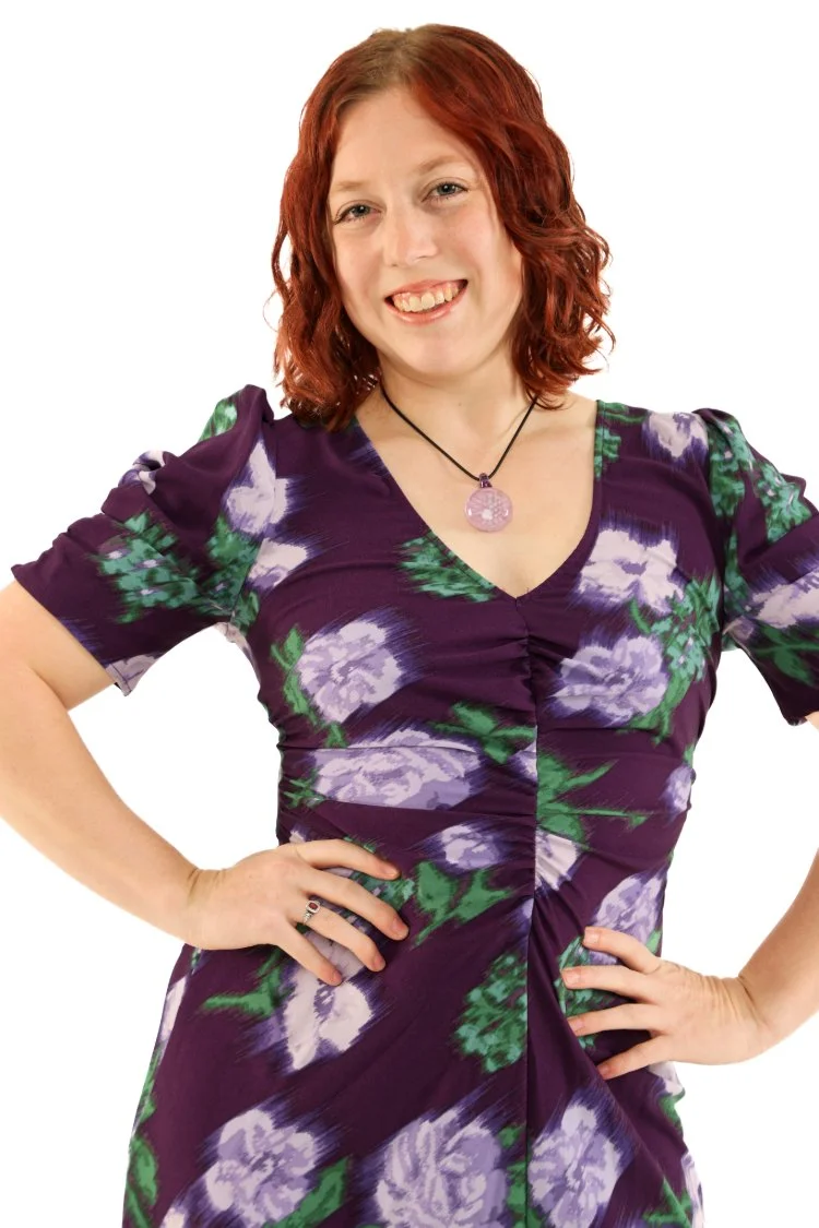 A woman with red curly hair is smiling and posing with hands on hips, wearing a purple floral dress and a purple pendant necklace.