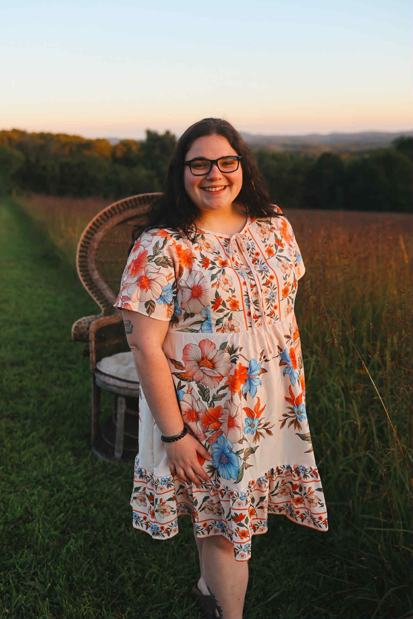 Smiling woman with dark hair wearing glasses, a red sweater, and ripped jeans, sitting beside a rustic wooden structure.