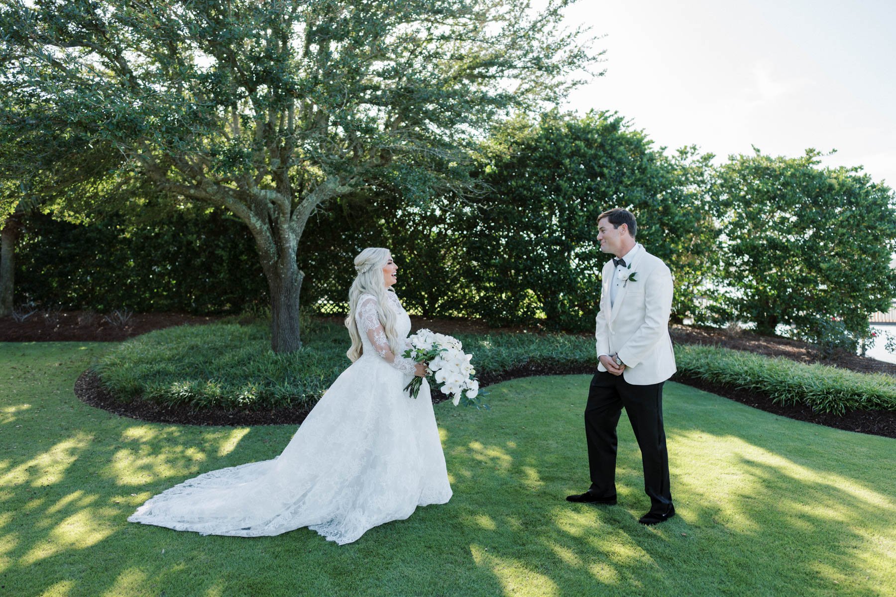 A wedding couple stands on a grassy area under a tree, with the bride holding a large bouquet of white flowers, while the groom looks at her. The bride wears a white lace wedding dress with a long train, and the groom wears a white tuxedo jacket and 