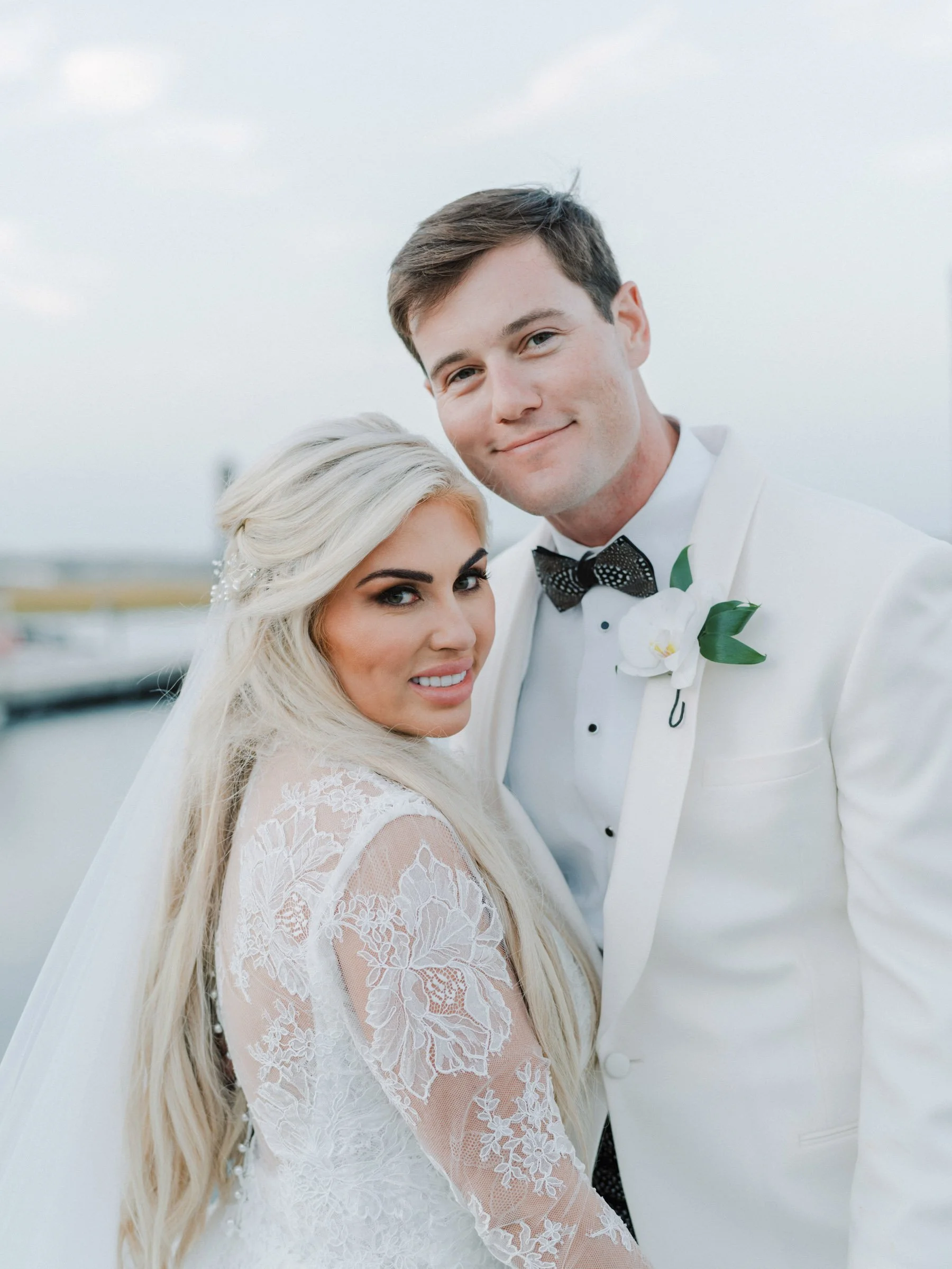 A bride and groom in wedding attire standing close together outdoors near a body of water.