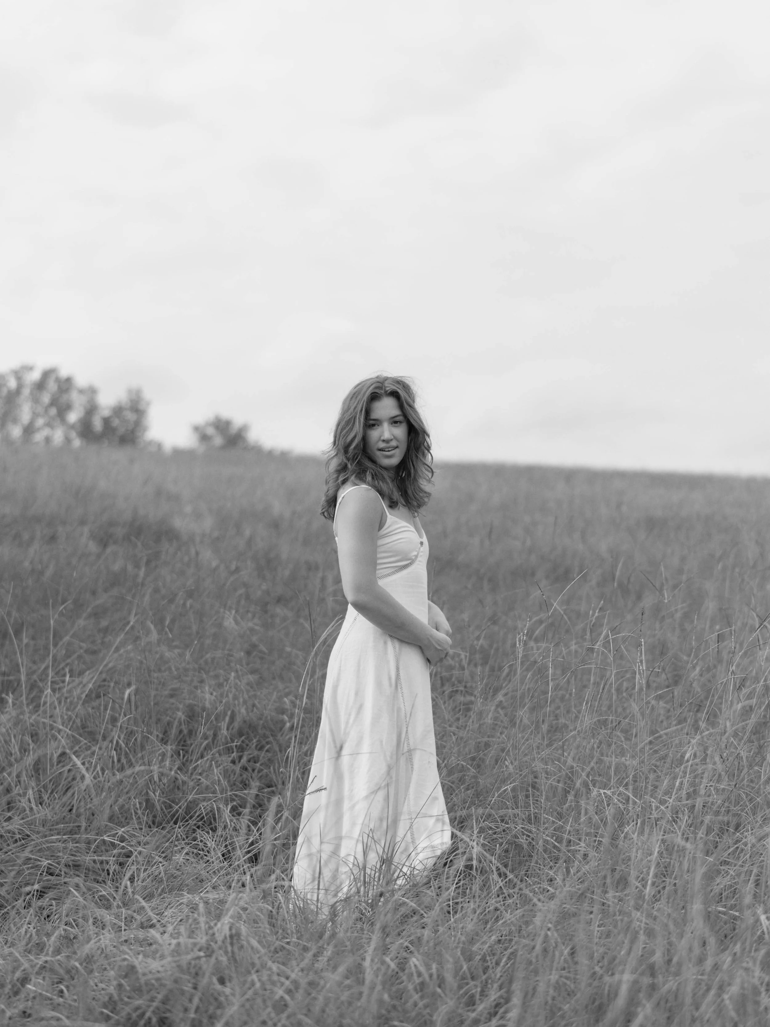 A woman wearing a light dress standing in a grassy field, looking at the camera, in black and white on her engagement session.